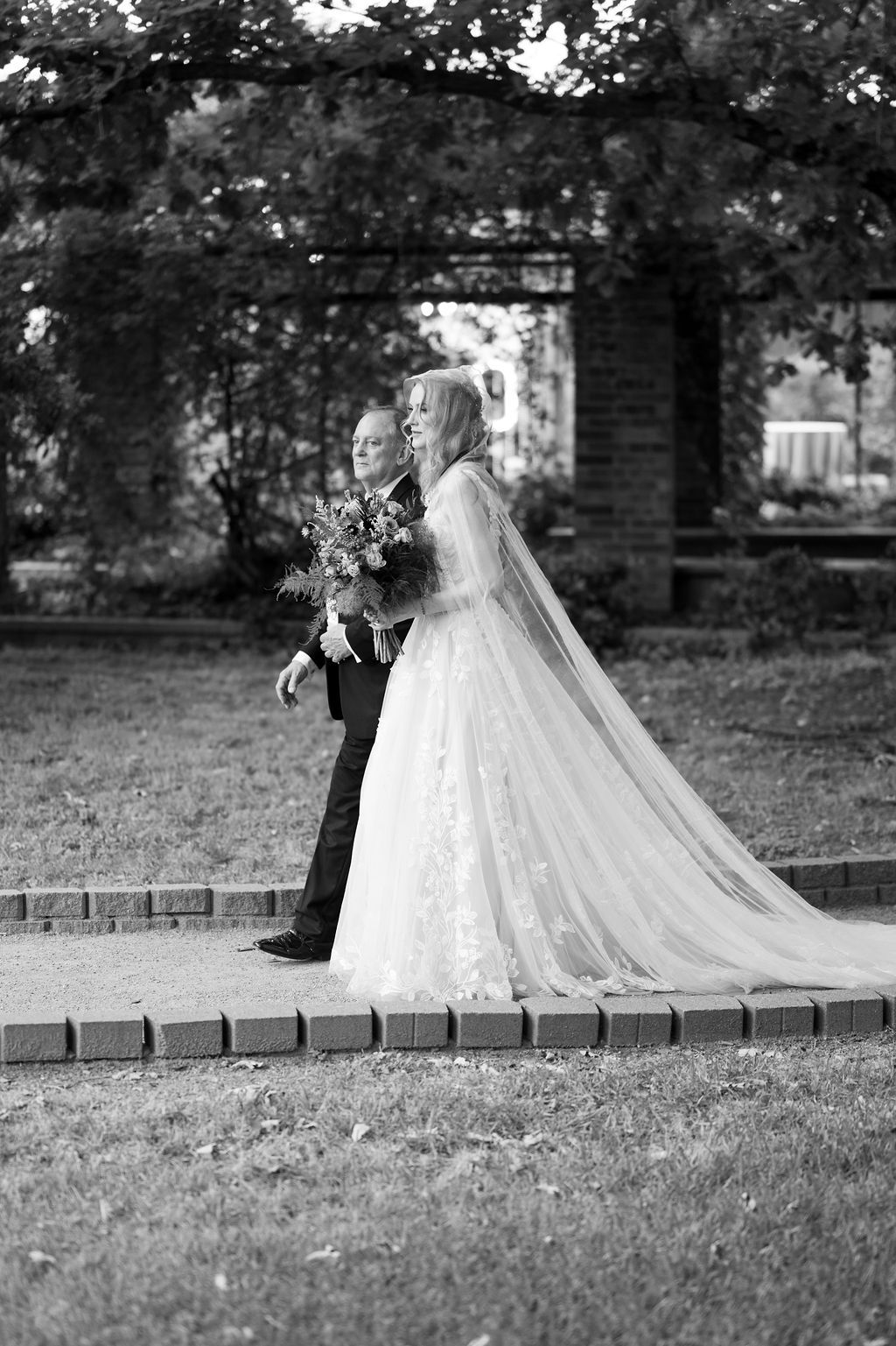 Black and white photo of bride in lace gown and veil walking with escort through garden arbor.