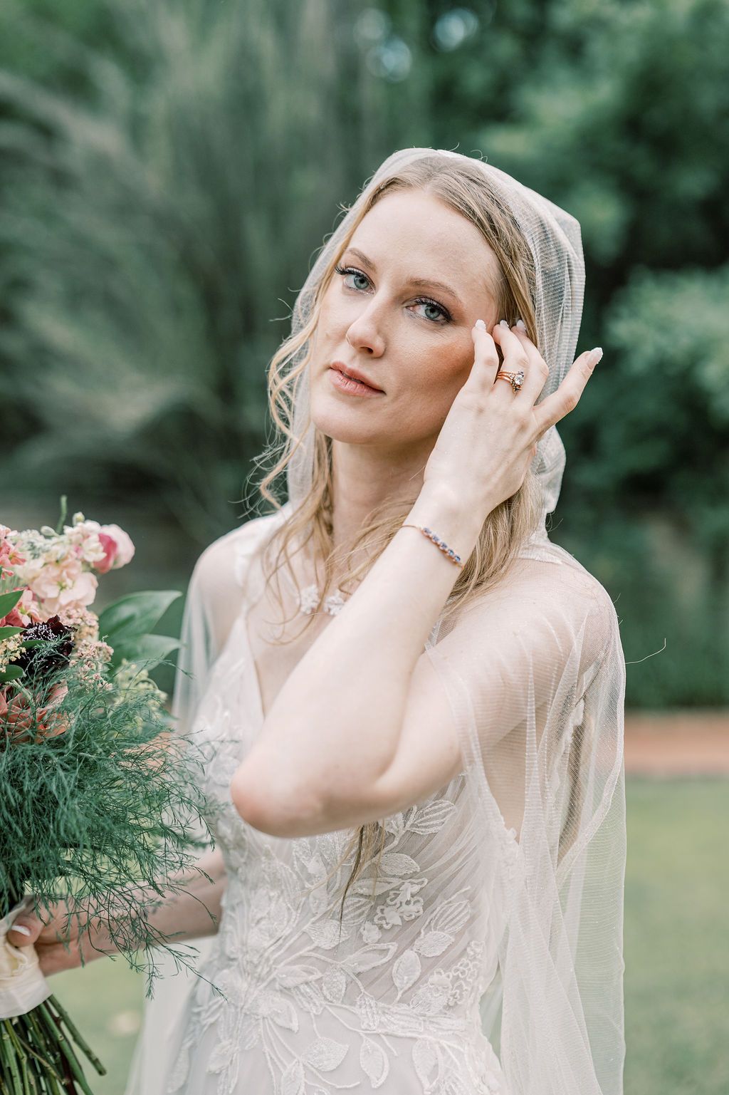 Blonde bride in lace gown and veil holds bouquet, posing outdoors among lush greenery.
