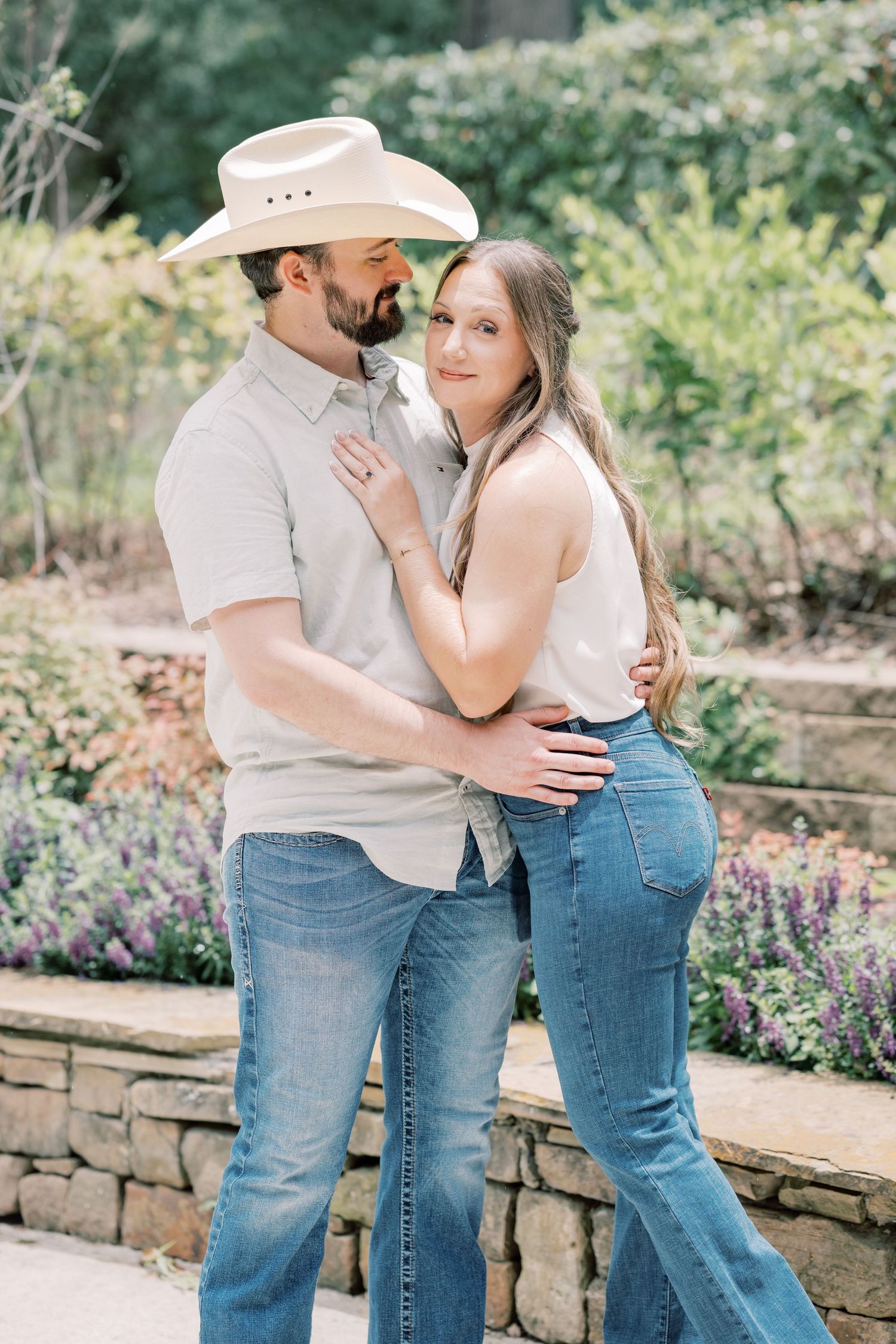 Couple embracing in a garden, man wearing cowboy hat and jeans, woman in white top and flared jeans.