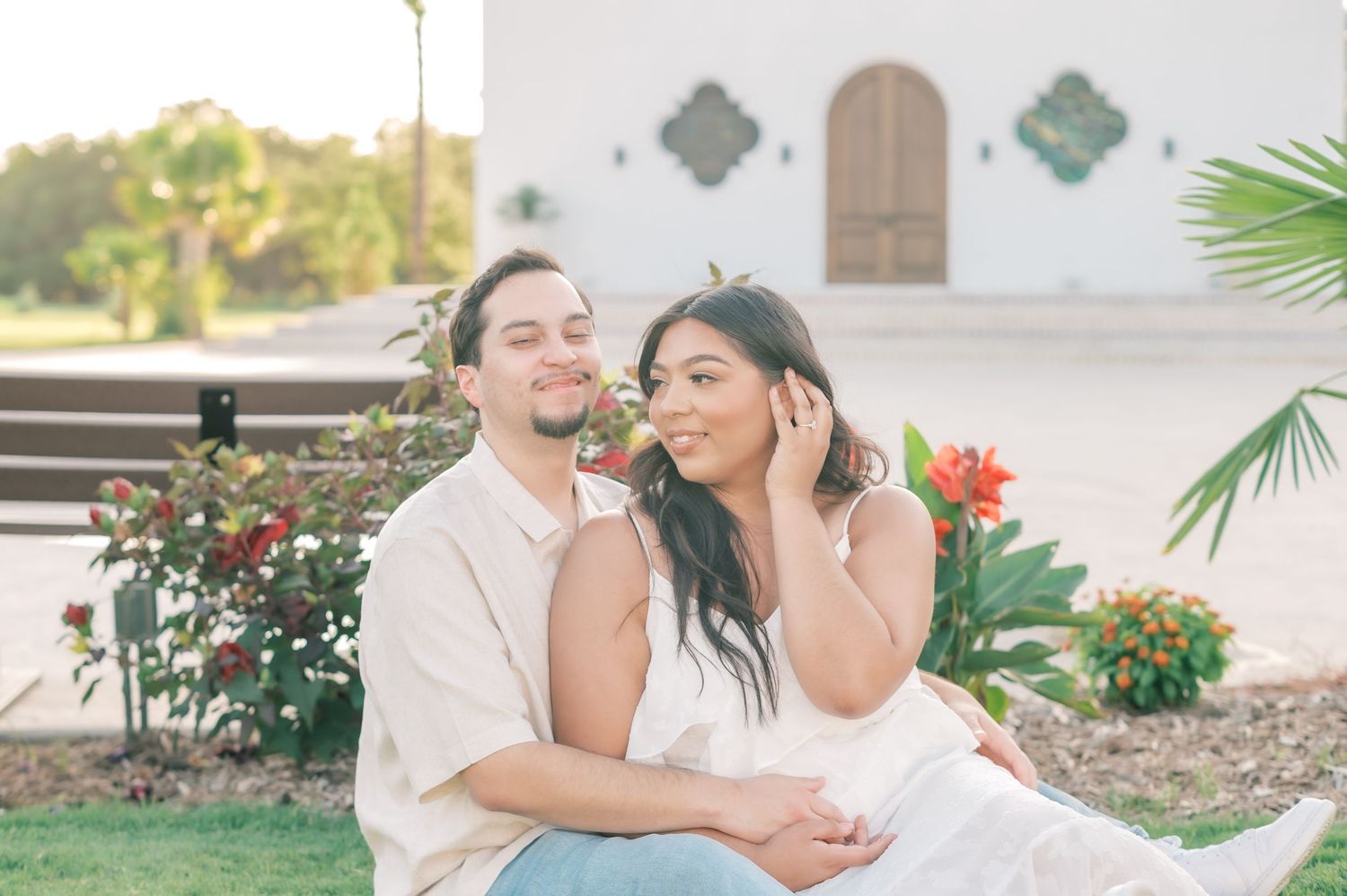 A couple embraces in a romantic outdoor garden setting with palm trees and flowers.