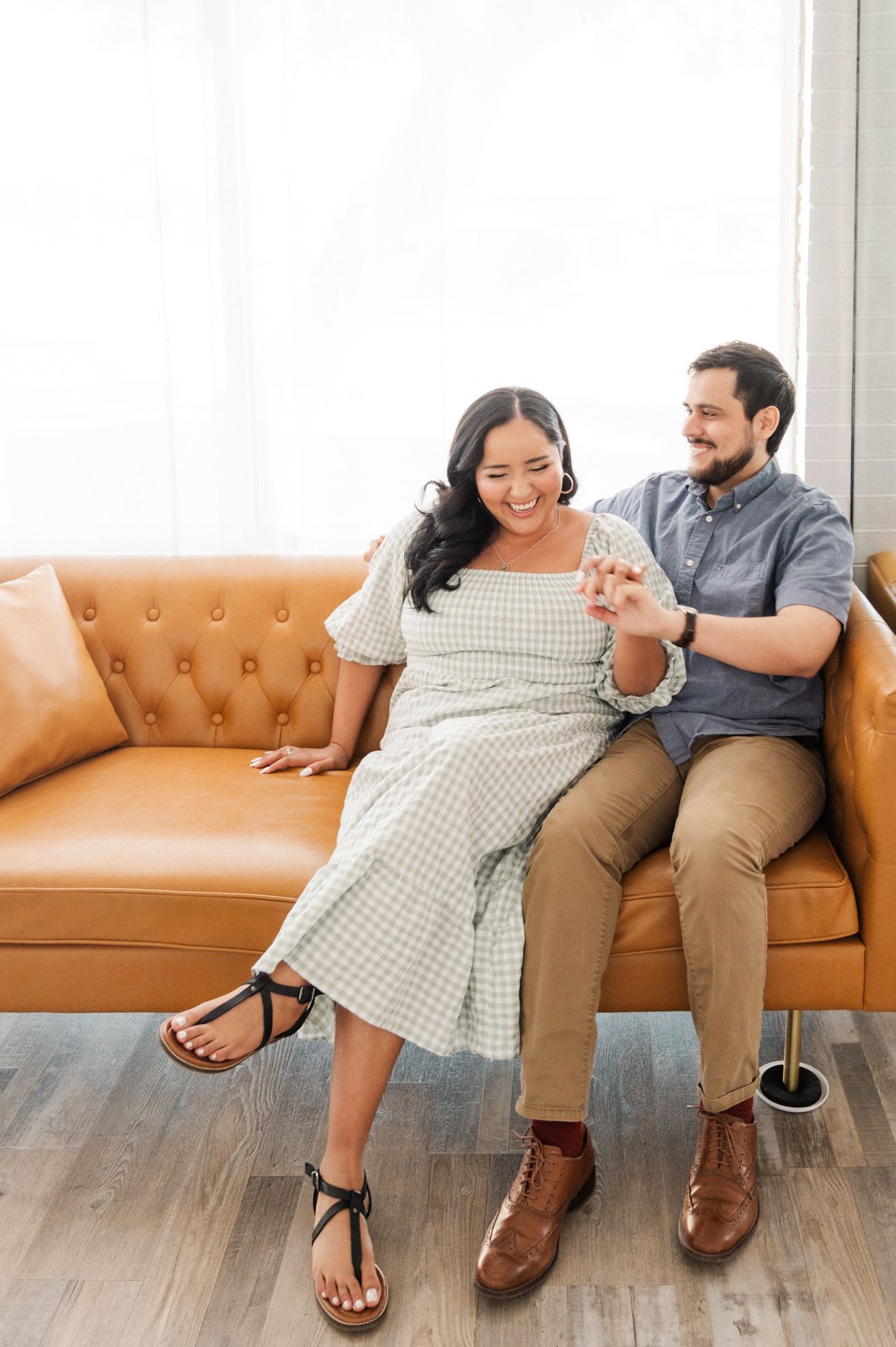A couple laughing together on a tan leather sofa in a sunlit room.