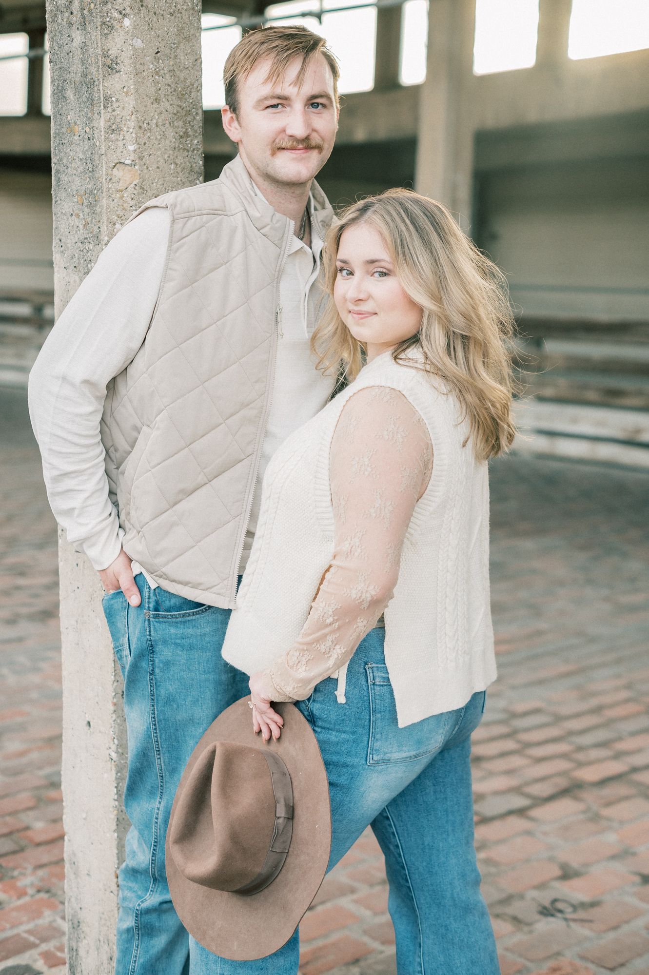 A couple in casual attire share a romantic moment while holding a brown cowboy hat in an industrial setting.