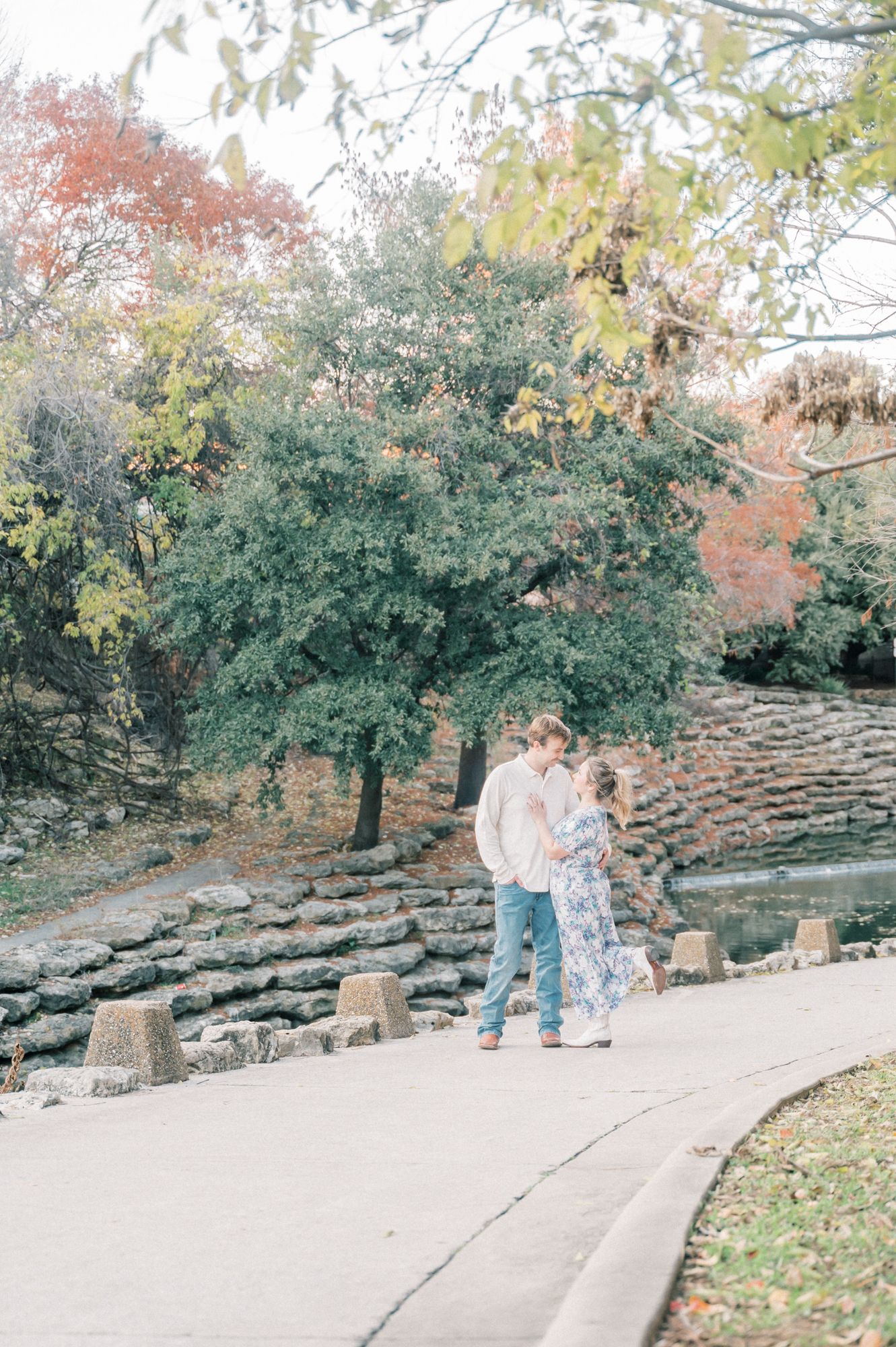 A couple stands on a paved pathway surrounded by autumn trees and stone terraces in a scenic park setting.