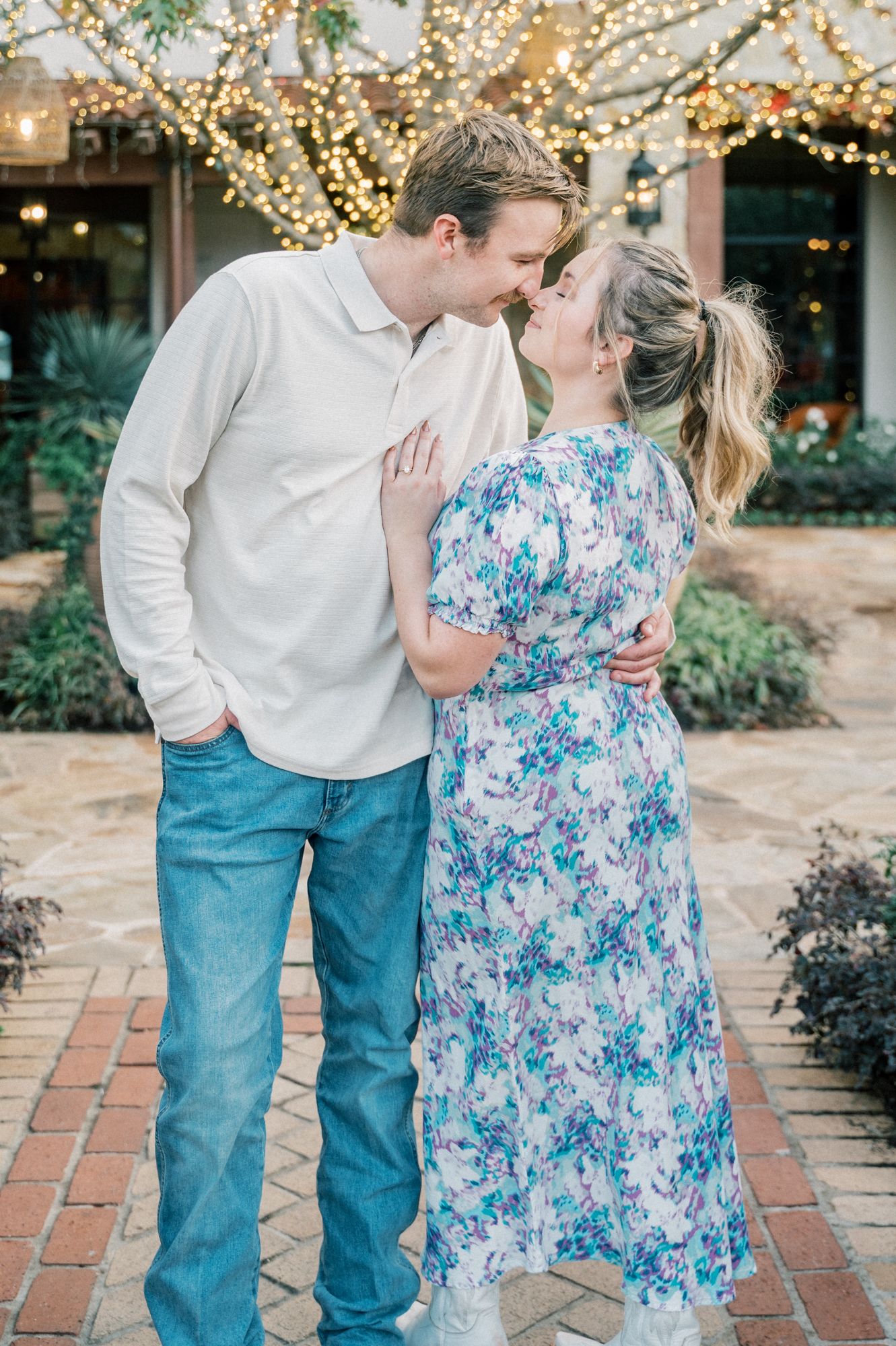 A couple in a floral dress and white shirt share romantic moments on a brick pathway with twinkling lights overhead.