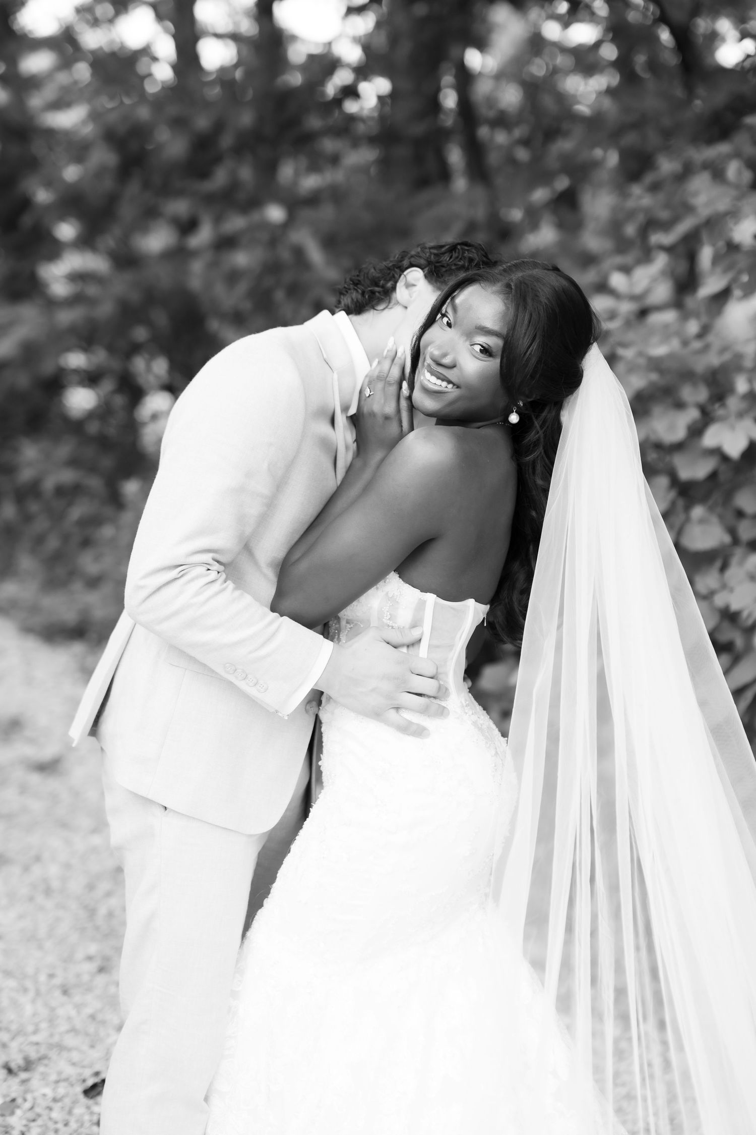Black and white photo of groom kissing bride on cheek, bride smiling in strapless lace wedding gown with long veil.
