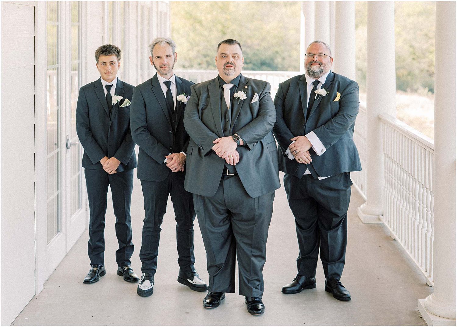 Groomsmen in dark suits pose together on a white columned porch.