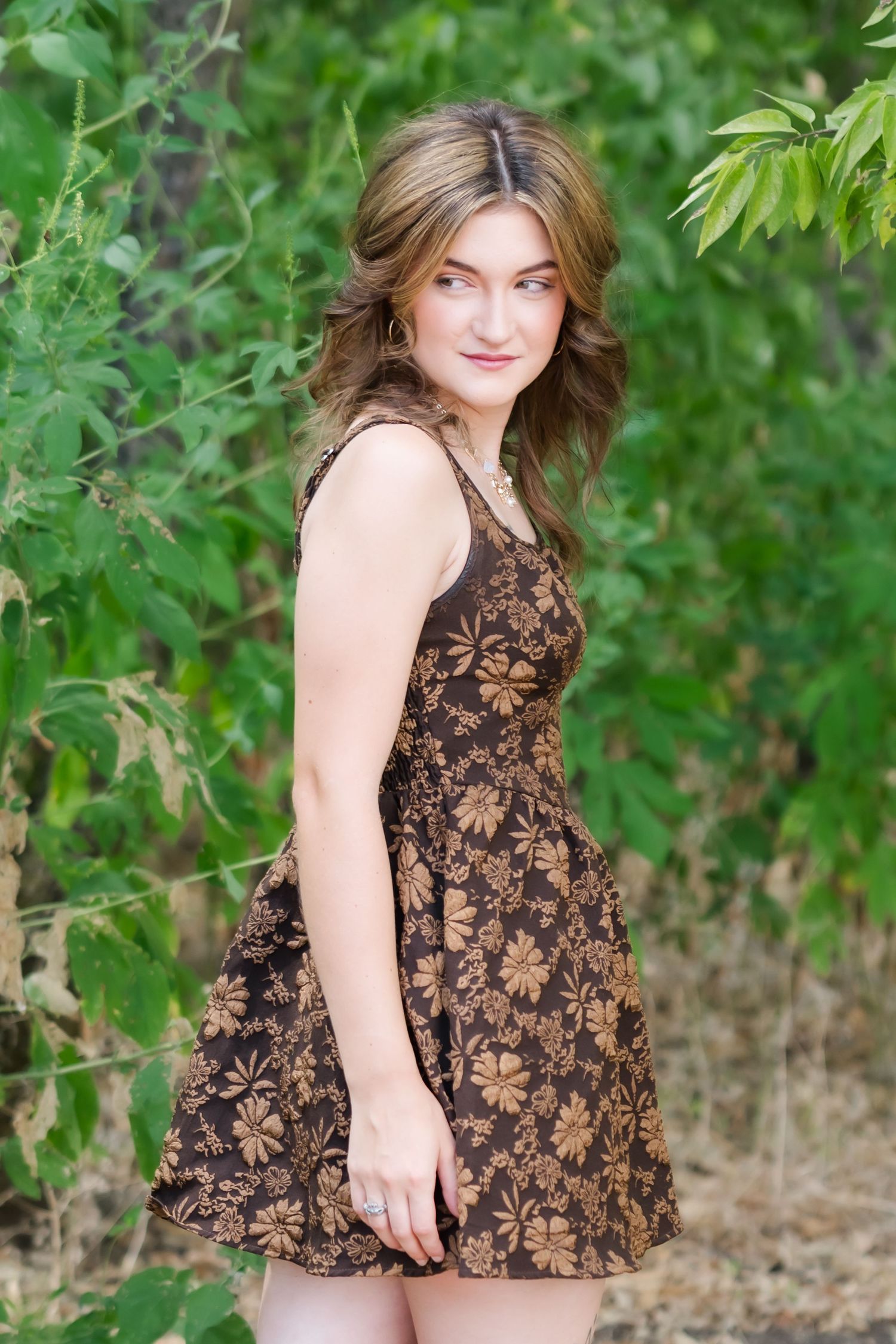 Young woman in brown floral sundress posing outdoors among lush green foliage, smiling naturally at camera.