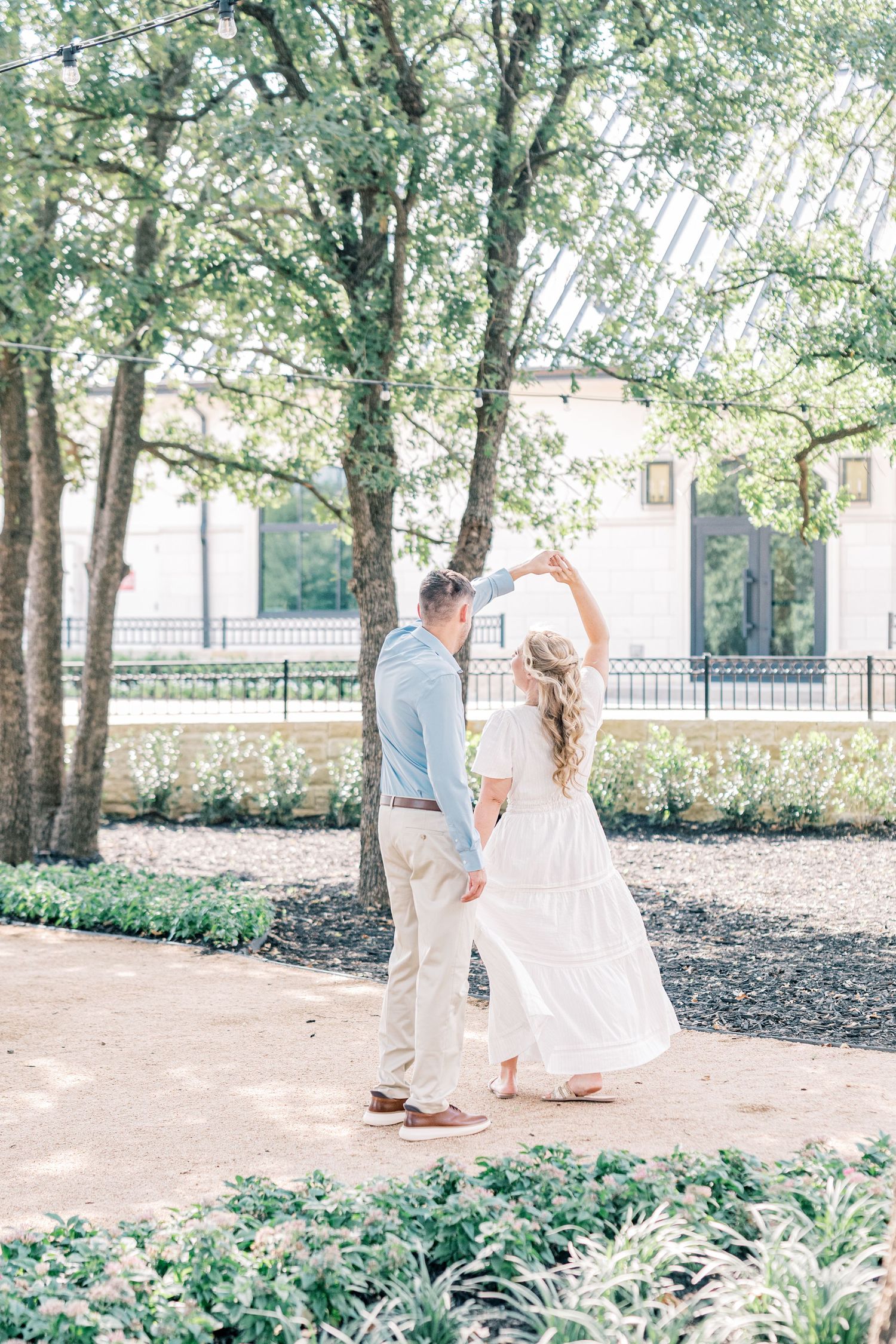 Couple dances outdoors in a sunlit garden, man twirling woman in a flowing white dress under trees.