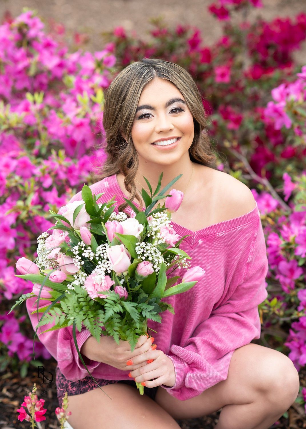 Smiling young woman in pink velvet sweater cradling floral bouquet while sitting among vibrant pink azalea bushes.