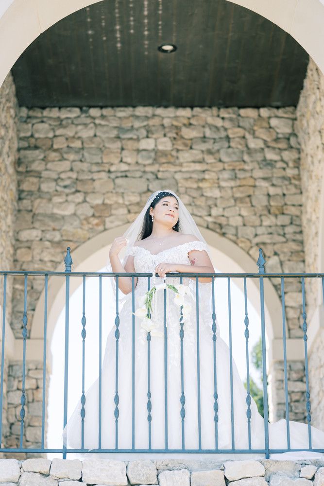 A bride in an off-shoulder white gown stands on a stone balcony with ornate metal railings.