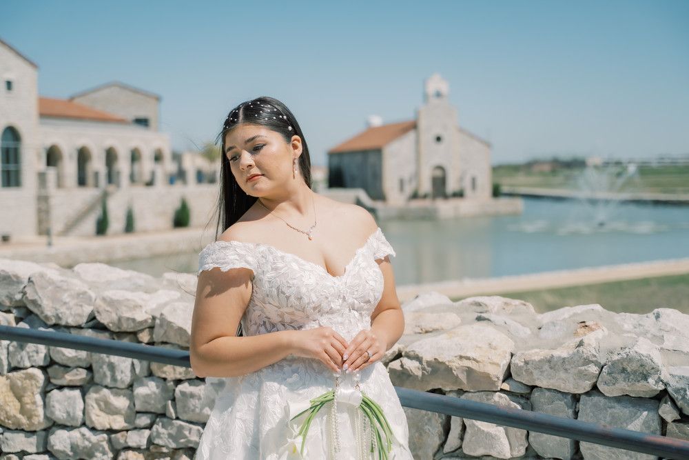 A coastal photoshoot features someone in a white dress posing gracefully by the ocean with stone architecture in view.