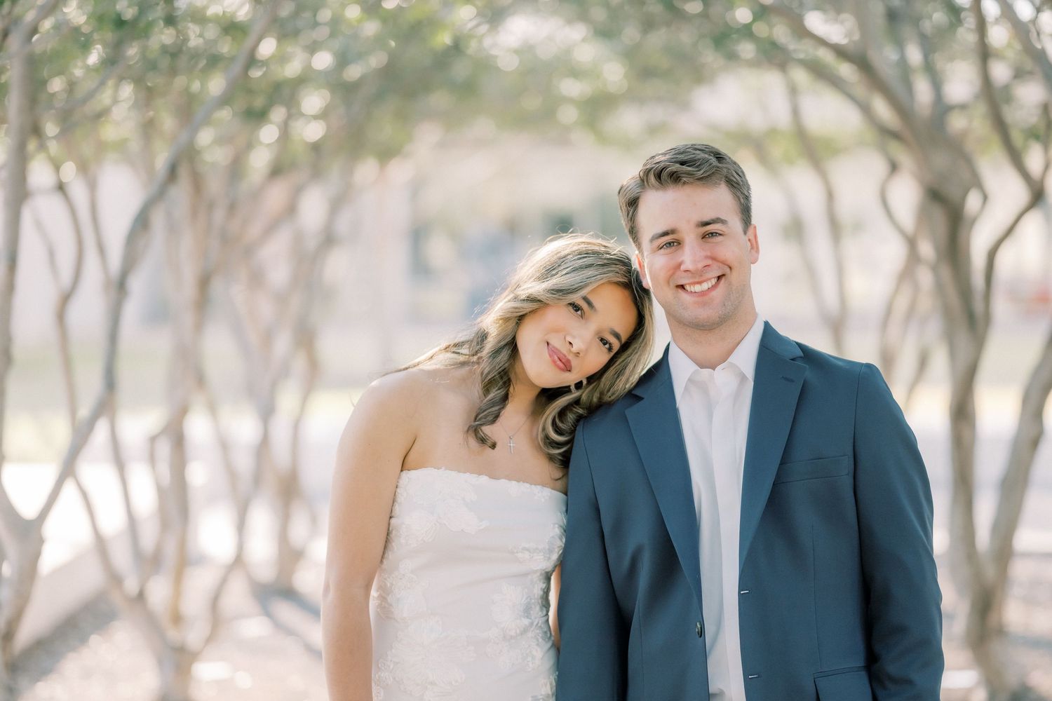 A couple in wedding attire embraces outdoors in a sun-dappled wooded setting.