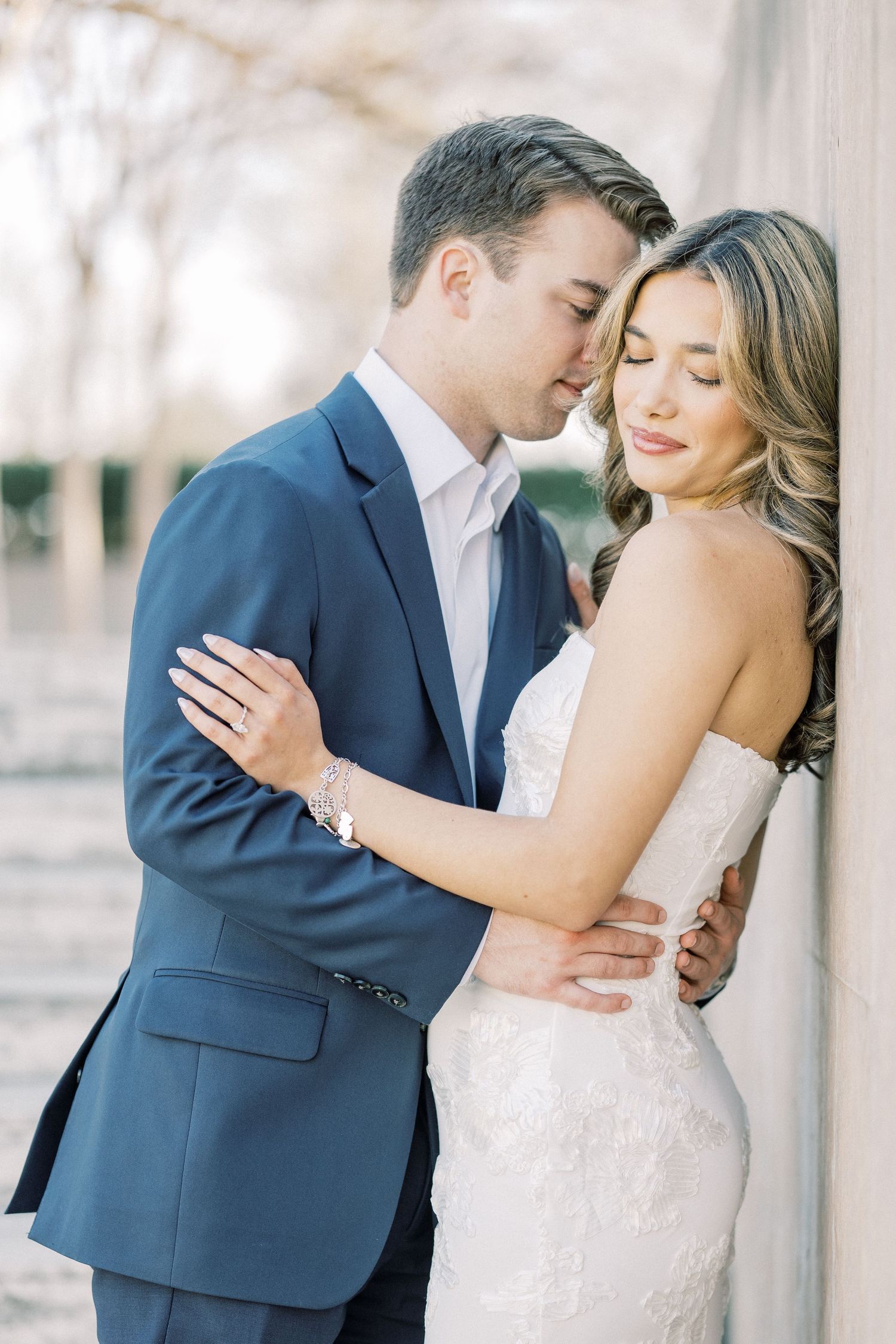 A romantic wedding couple embraces in front of white pillars wearing navy suit and white lace gown.