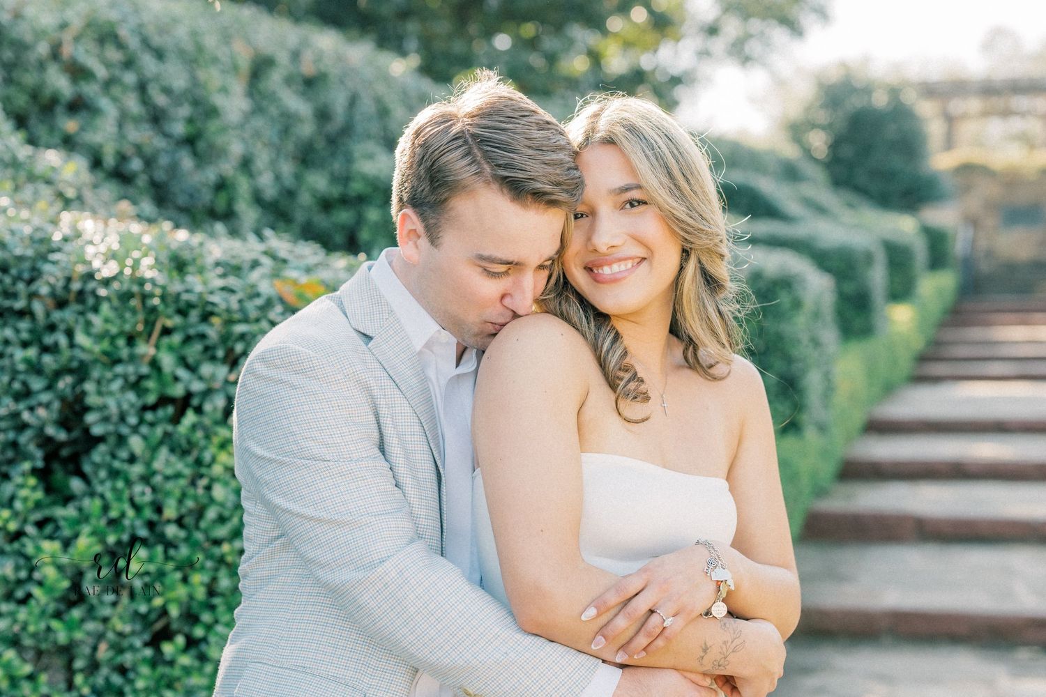 A couple embraces in a romantic garden setting with manicured hedges and stone steps in soft sunlight.
