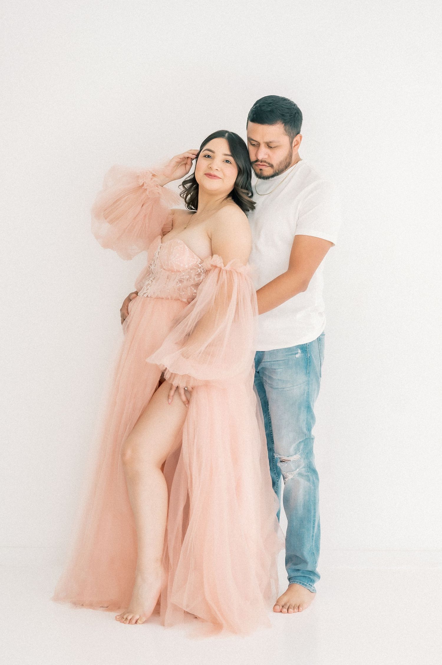 Expecting couple in a bright studio, woman in pink tulle gown, man in white tee and jeans.