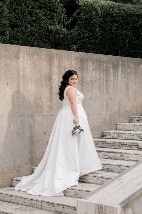 Bride in white ball gown holding bouquet poses on stone steps with lush green hedges in background.