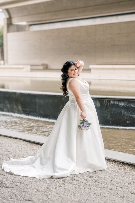 Bride in white ball gown with train holds bouquet near fountain.