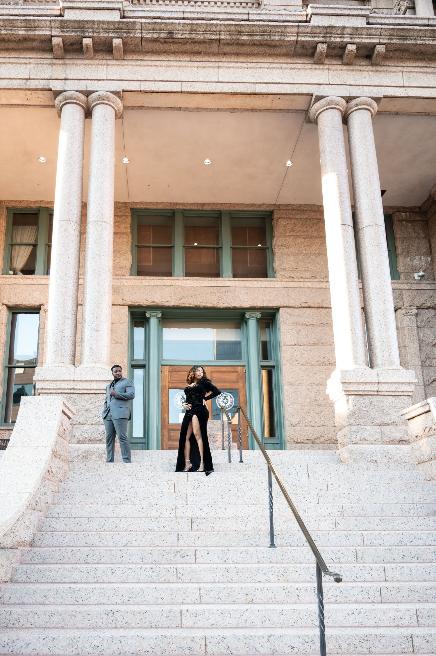 Couple posing on grand stone steps of a neoclassical building with tall columns, dressed in elegant formal attire.