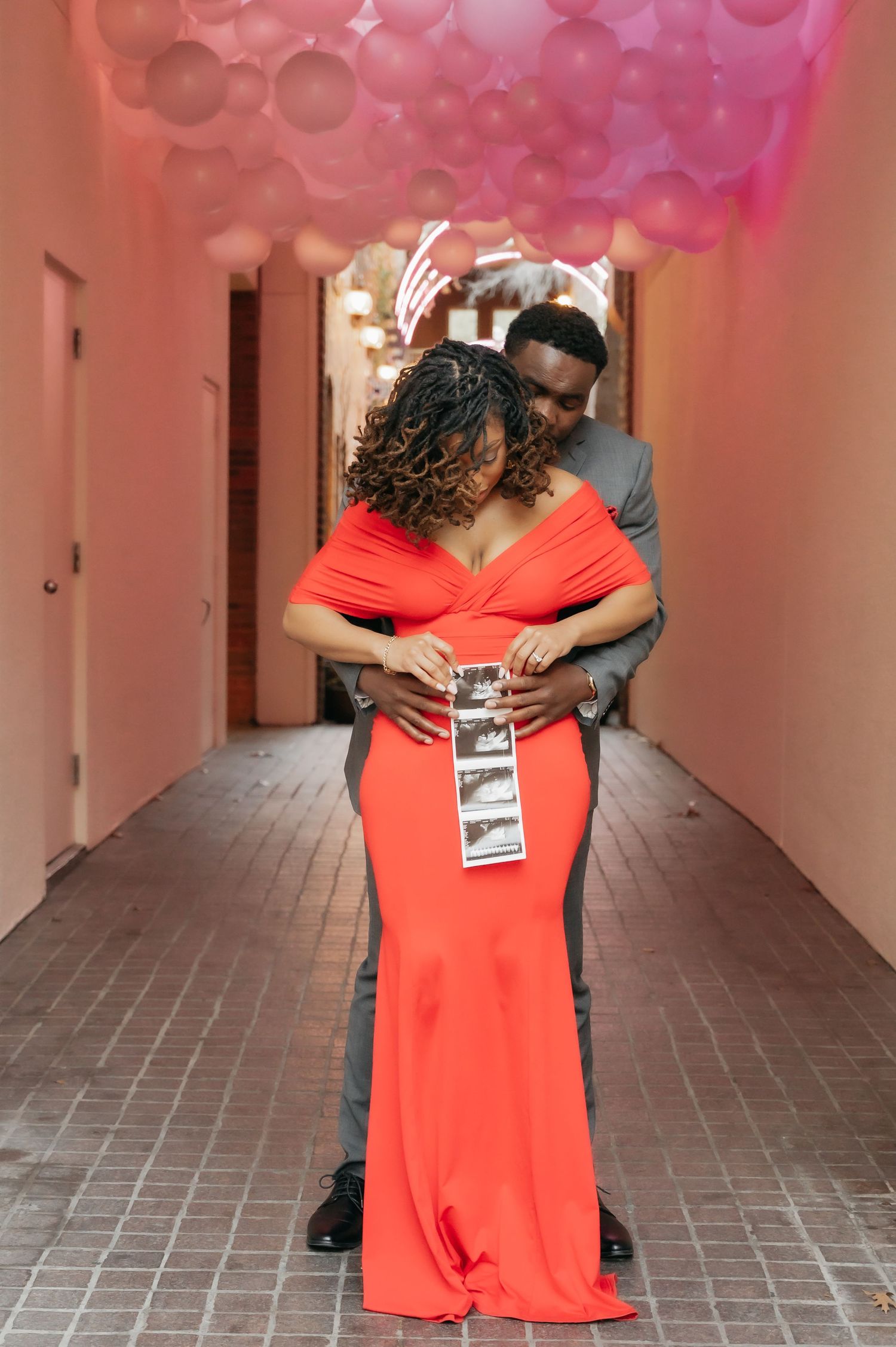 Couple in red gown holding ultrasound photos in pink balloon-decorated hallway during maternity announcement shoot.