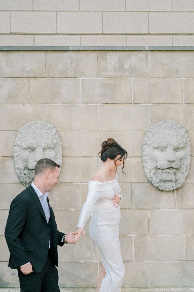 A couple in wedding attire holds hands walking past ornate lion head wall fountains on a stone building exterior.