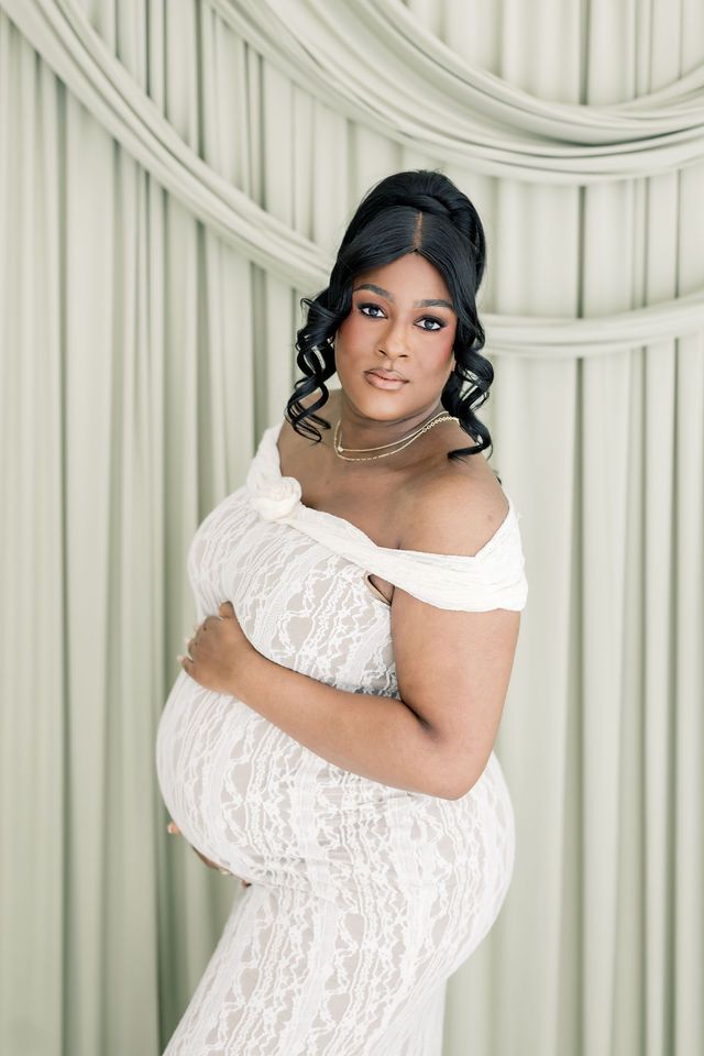 Elegant pregnant woman in white lace off-shoulder gown poses gracefully against soft draped backdrop.