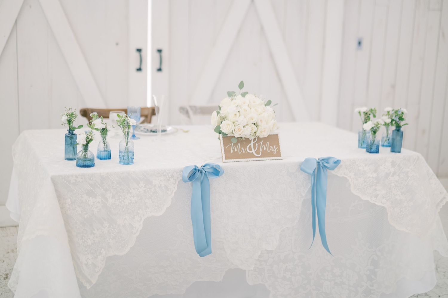 Sweetheart table with white lace cloth, blue ribbon ties, and white floral centerpiece in barn.