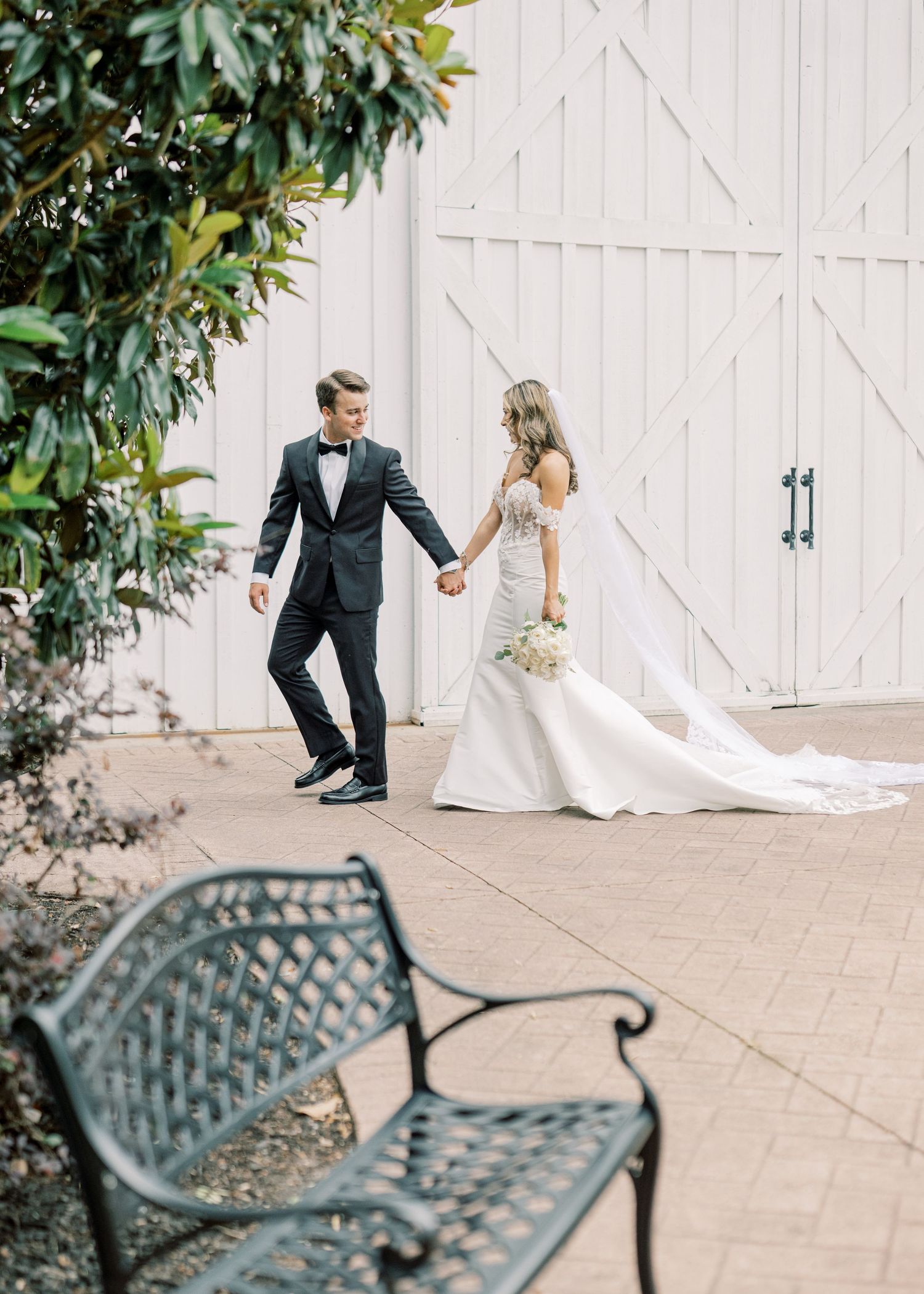 Bride and groom holding hands walking past white barn doors on brick patio on wedding day.