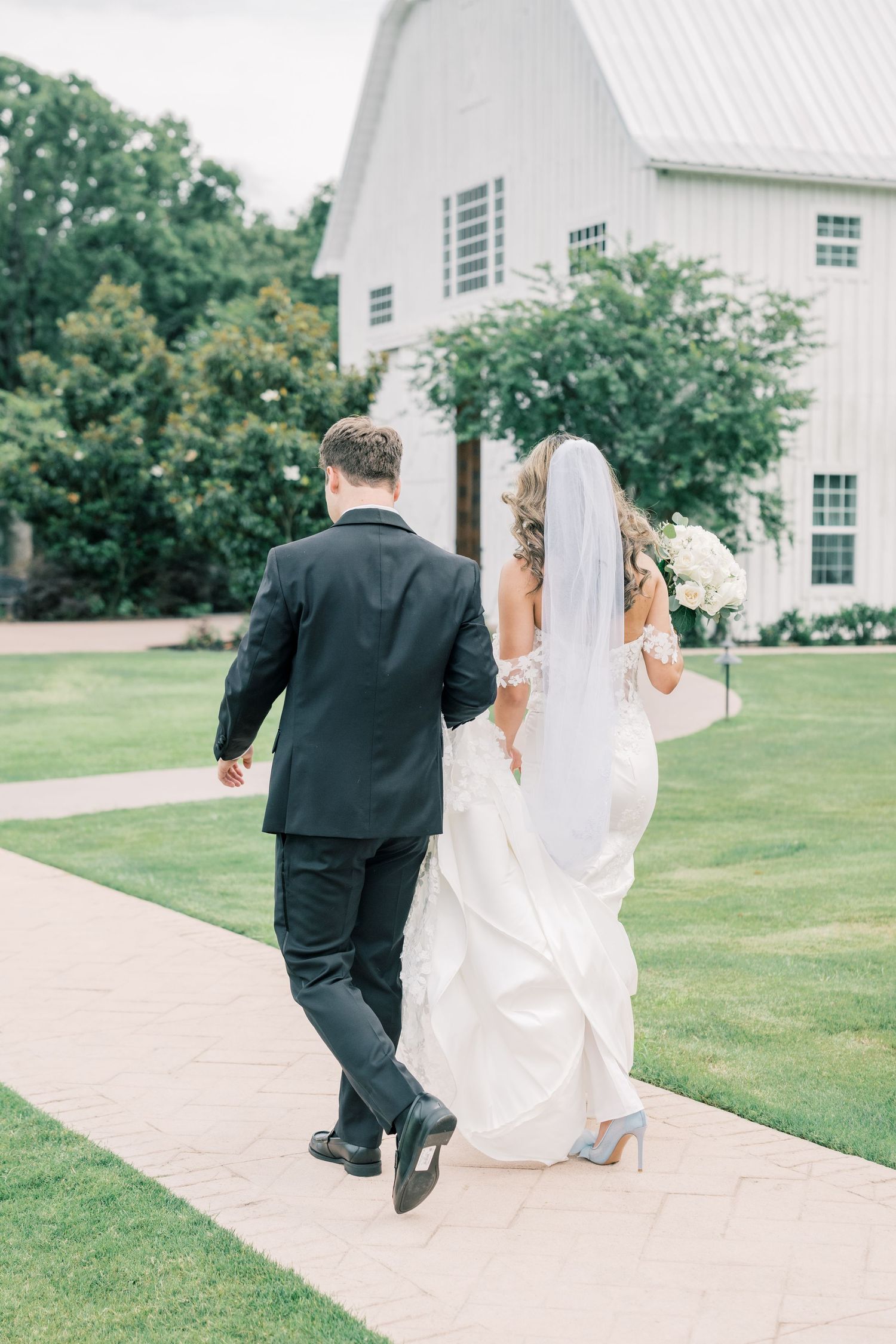 Bride and groom walking hand in hand toward white barn venue on manicured lawn path.