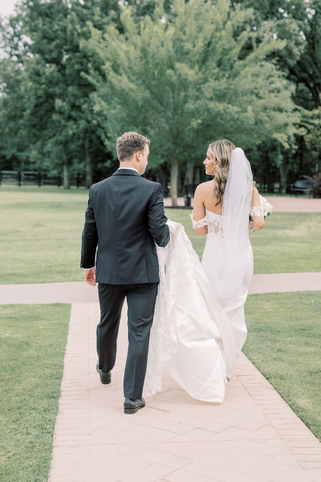 Bride and groom walk hand-in-hand down a garden path, bride in off-shoulder gown with veil, groom in black suit.