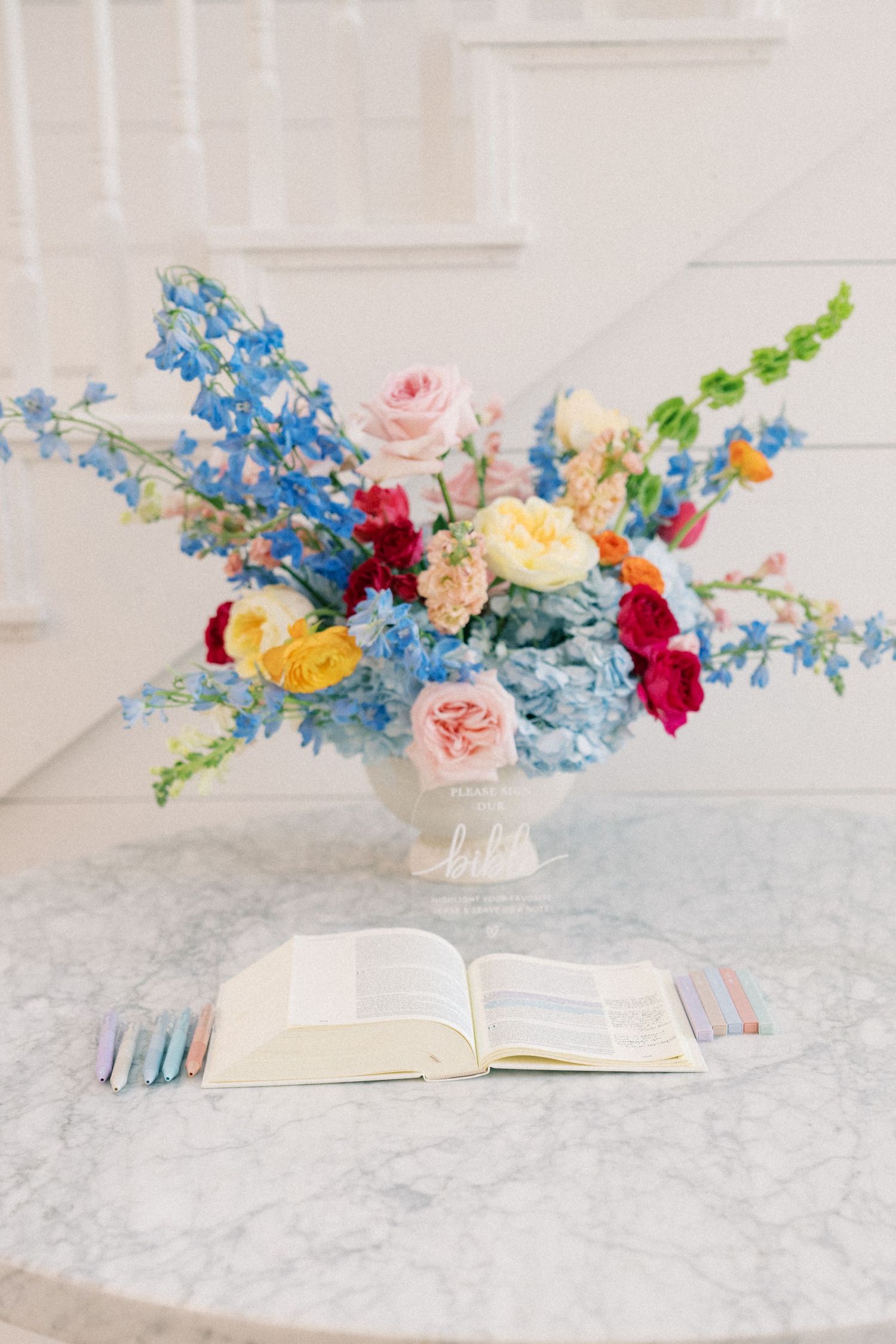 Colorful floral arrangement with blue hydrangeas, roses, and delphiniums in white vase on marble table with open book.