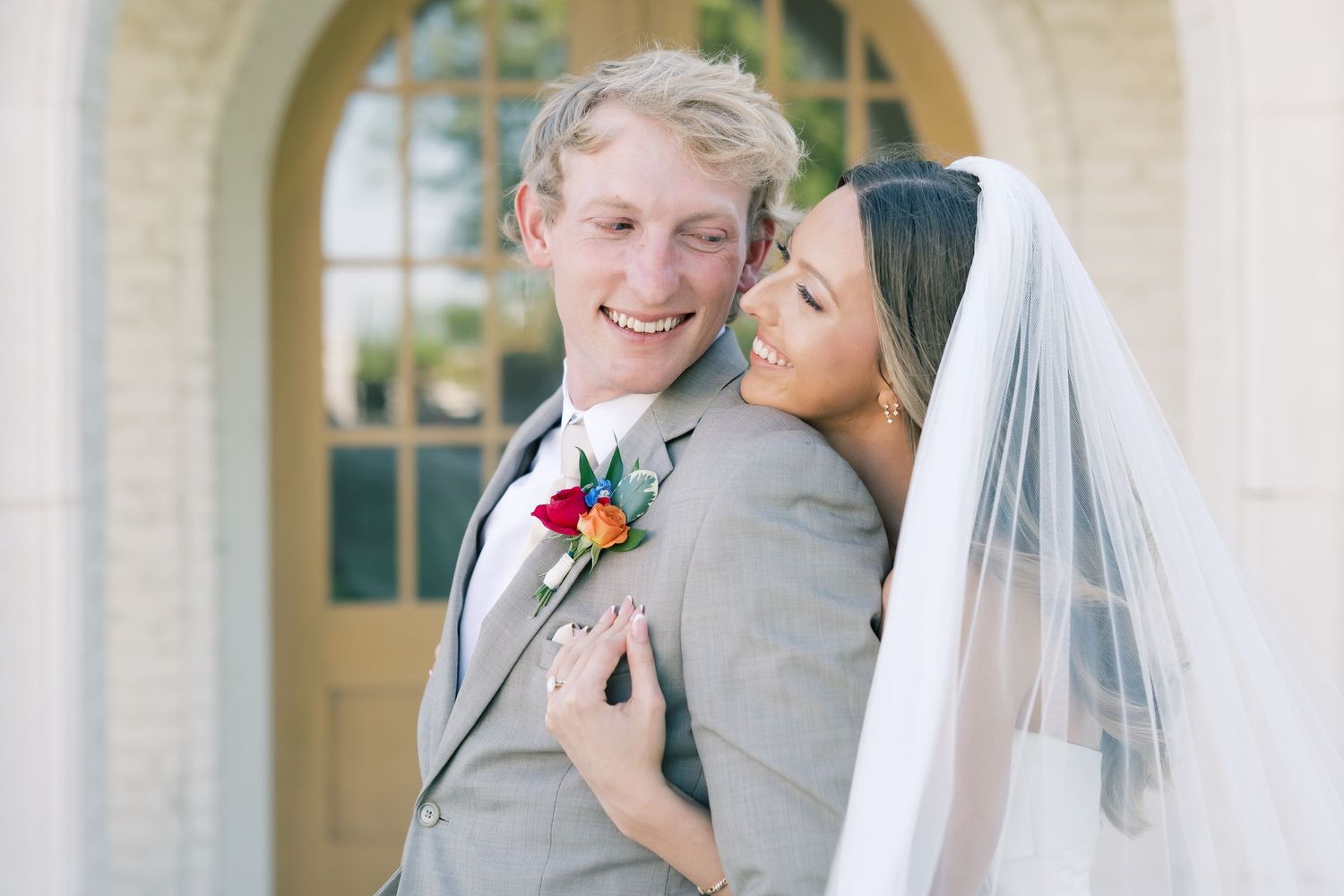 Happy newlyweds pose outside a church, bride in veil embracing groom in gray suit with colorful boutonniere.