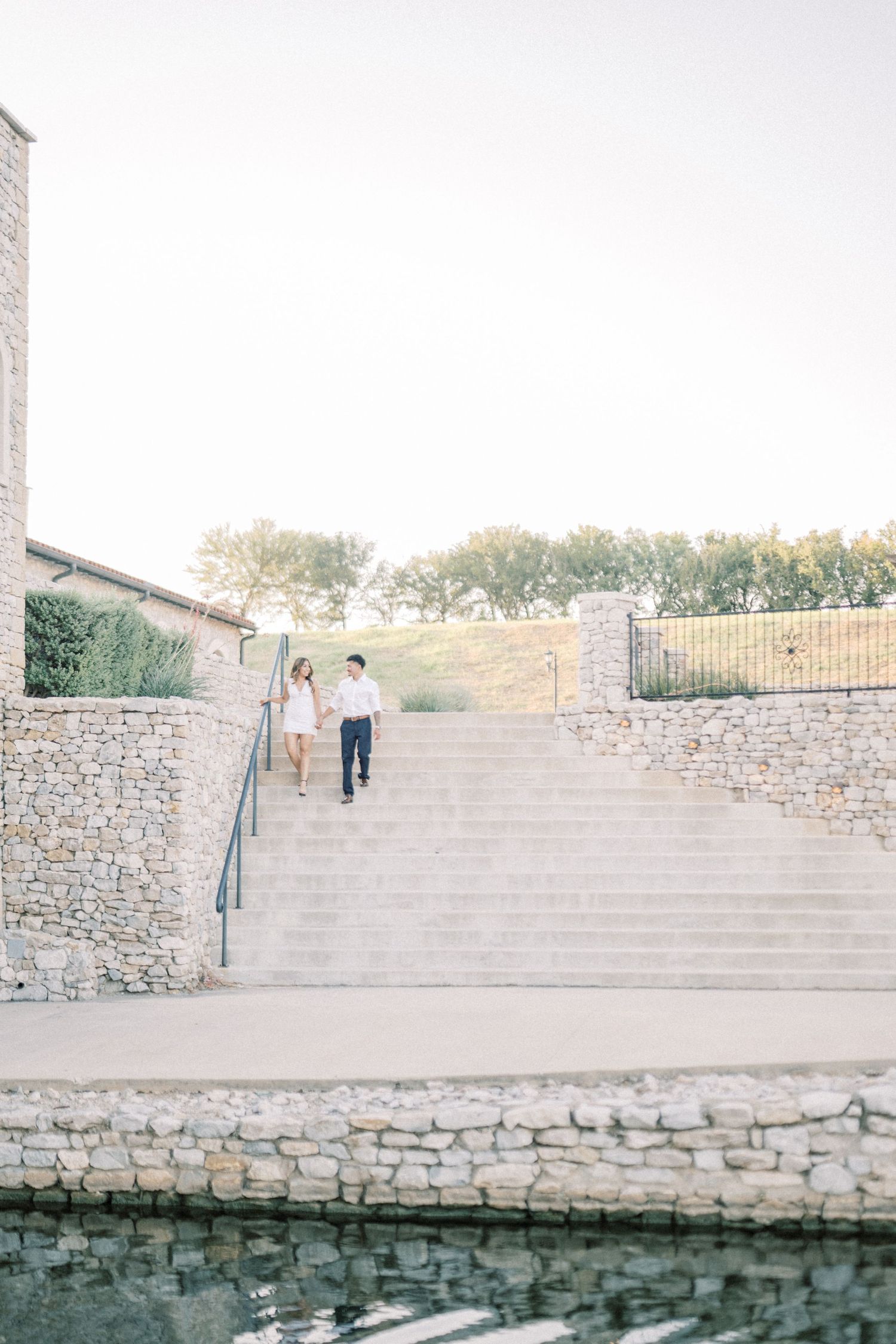 Engaged couple holding hands walking down stone steps near a reflecting pool at sunset.