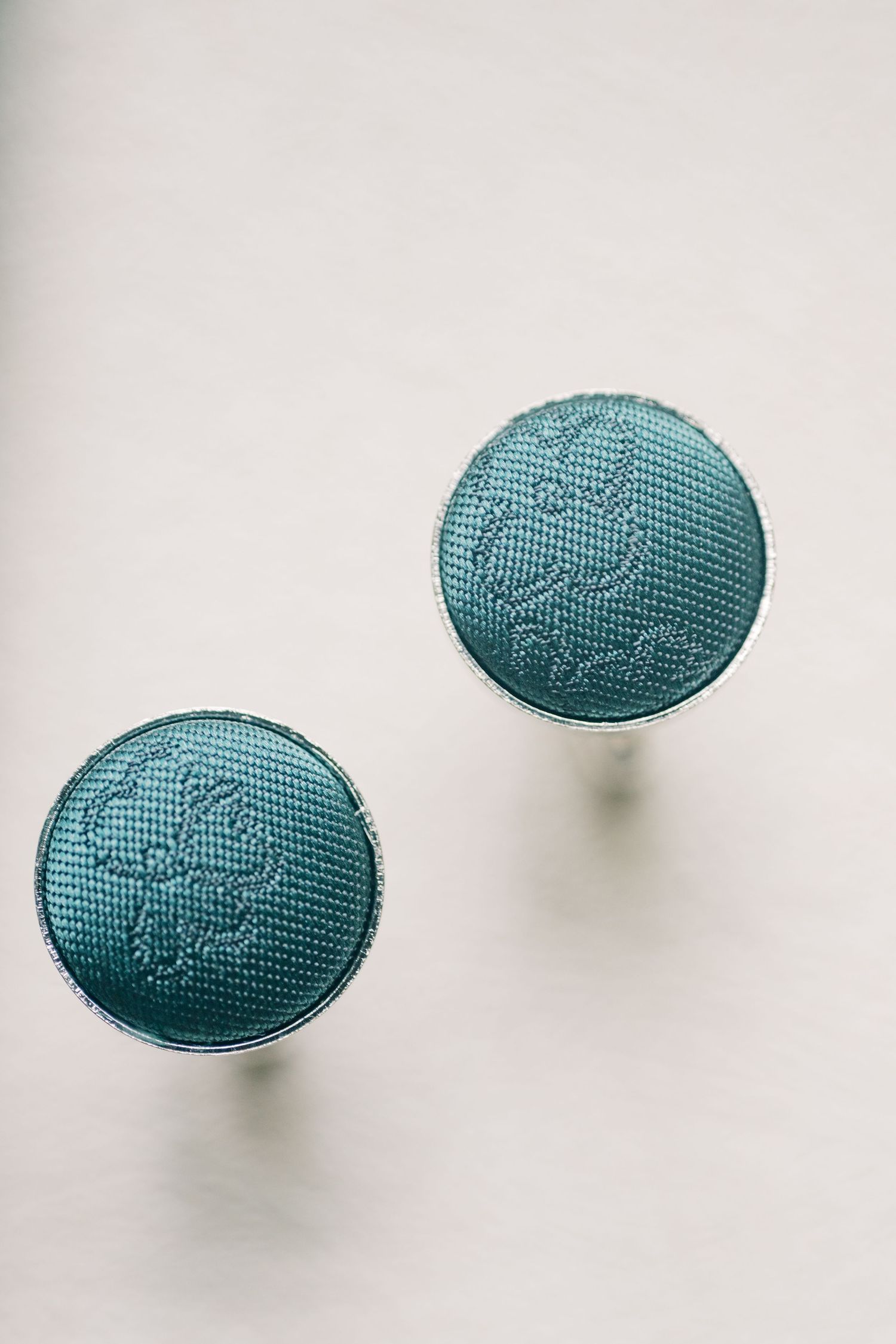 Teal fabric button cufflinks with floral embroidery pattern and silver edges on white background.