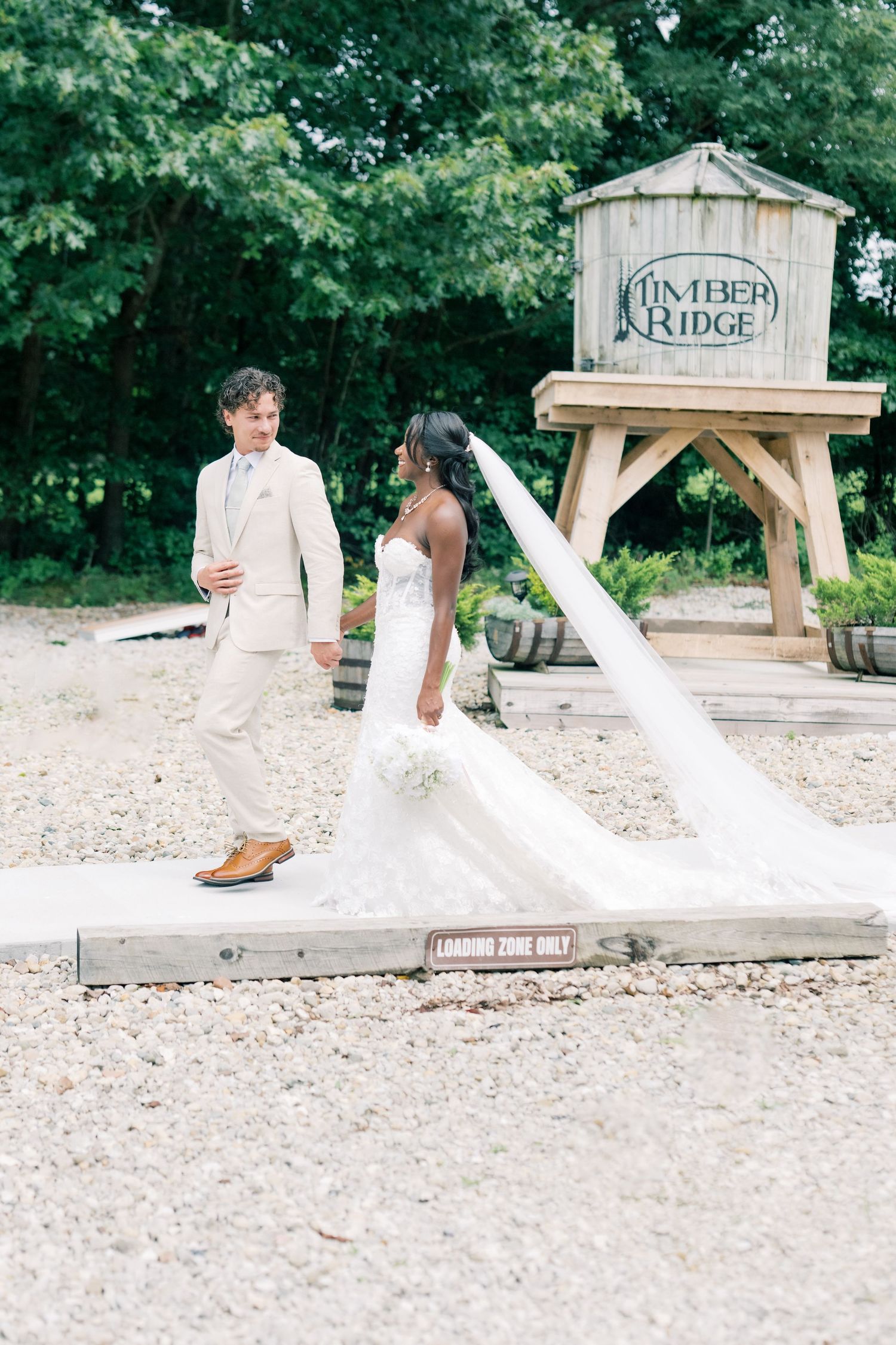 Interracial couple walks hand in hand at Timber Ridge venue, bride in white gown with flowing cathedral veil.