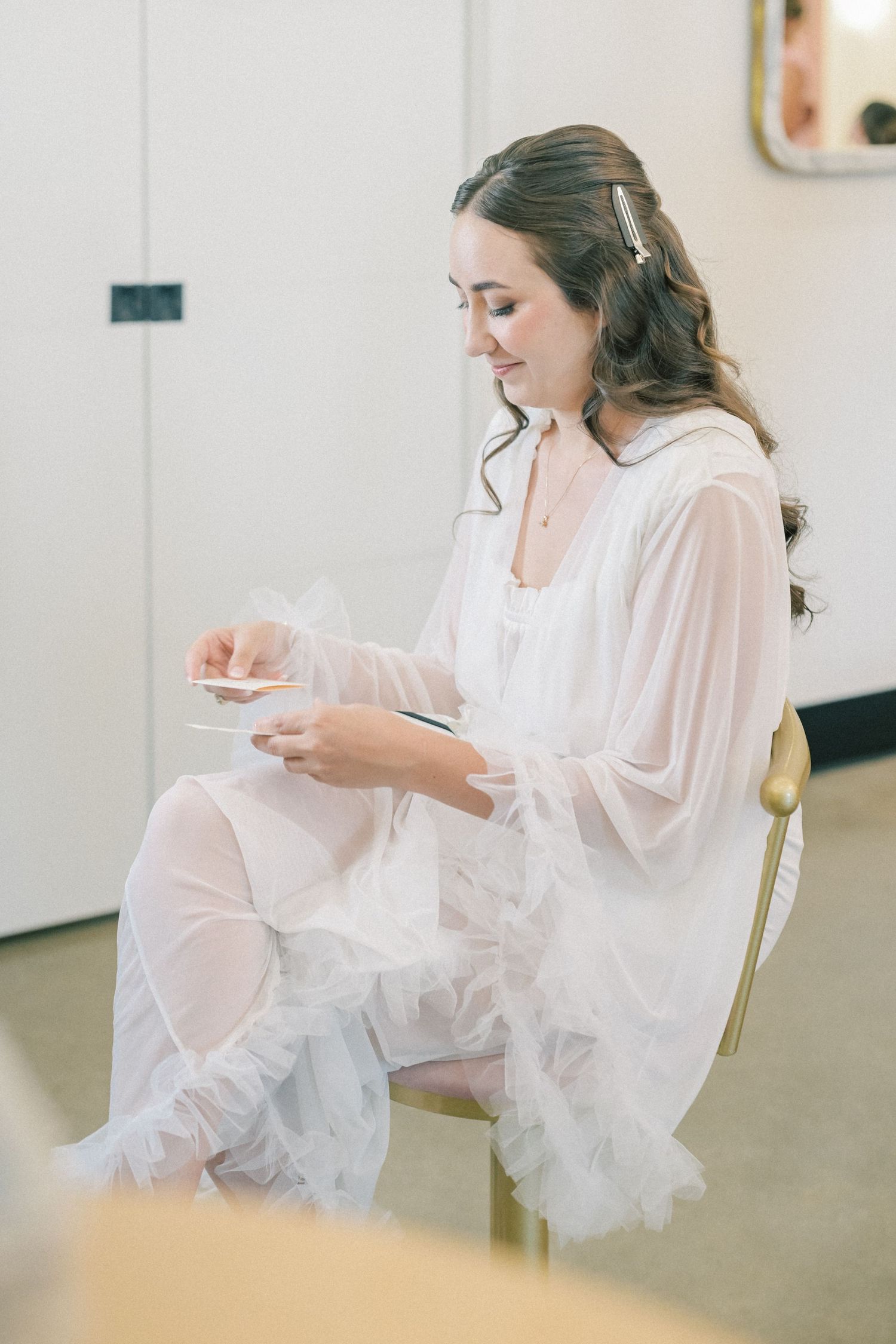 Bride in sheer white robe reading a note while seated in a gold chair during wedding preparations.