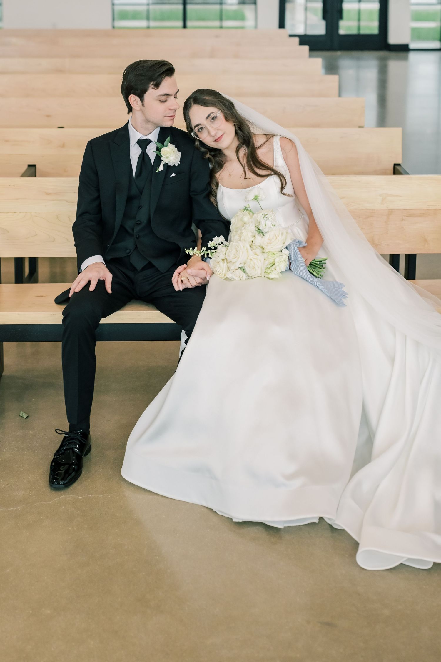 Bride and groom sit closely together on a wooden church pew, bride holding a white bouquet in her flowing wedding gown.