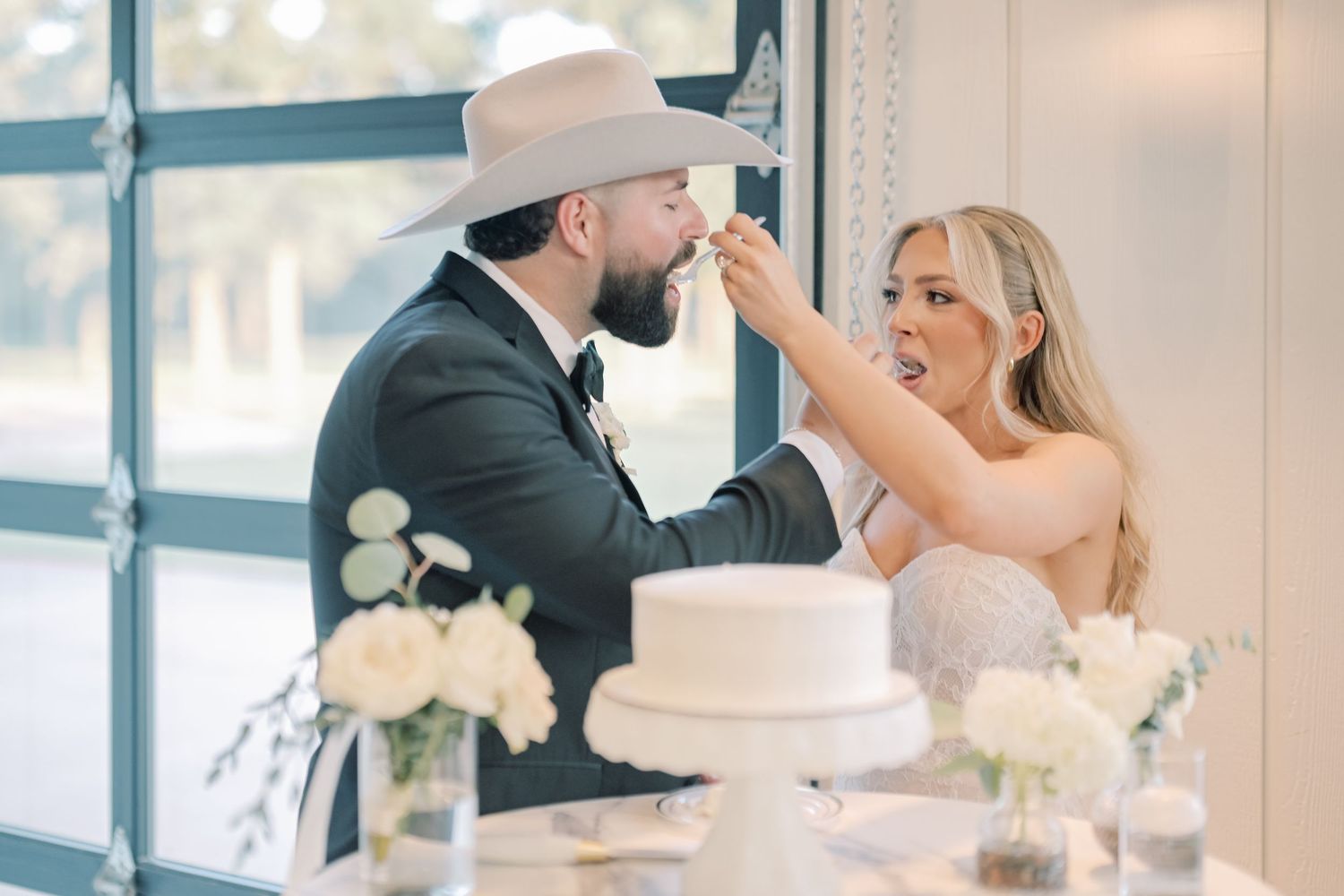 Bride feeds groom in cowboy hat wedding cake at white floral reception dessert table.