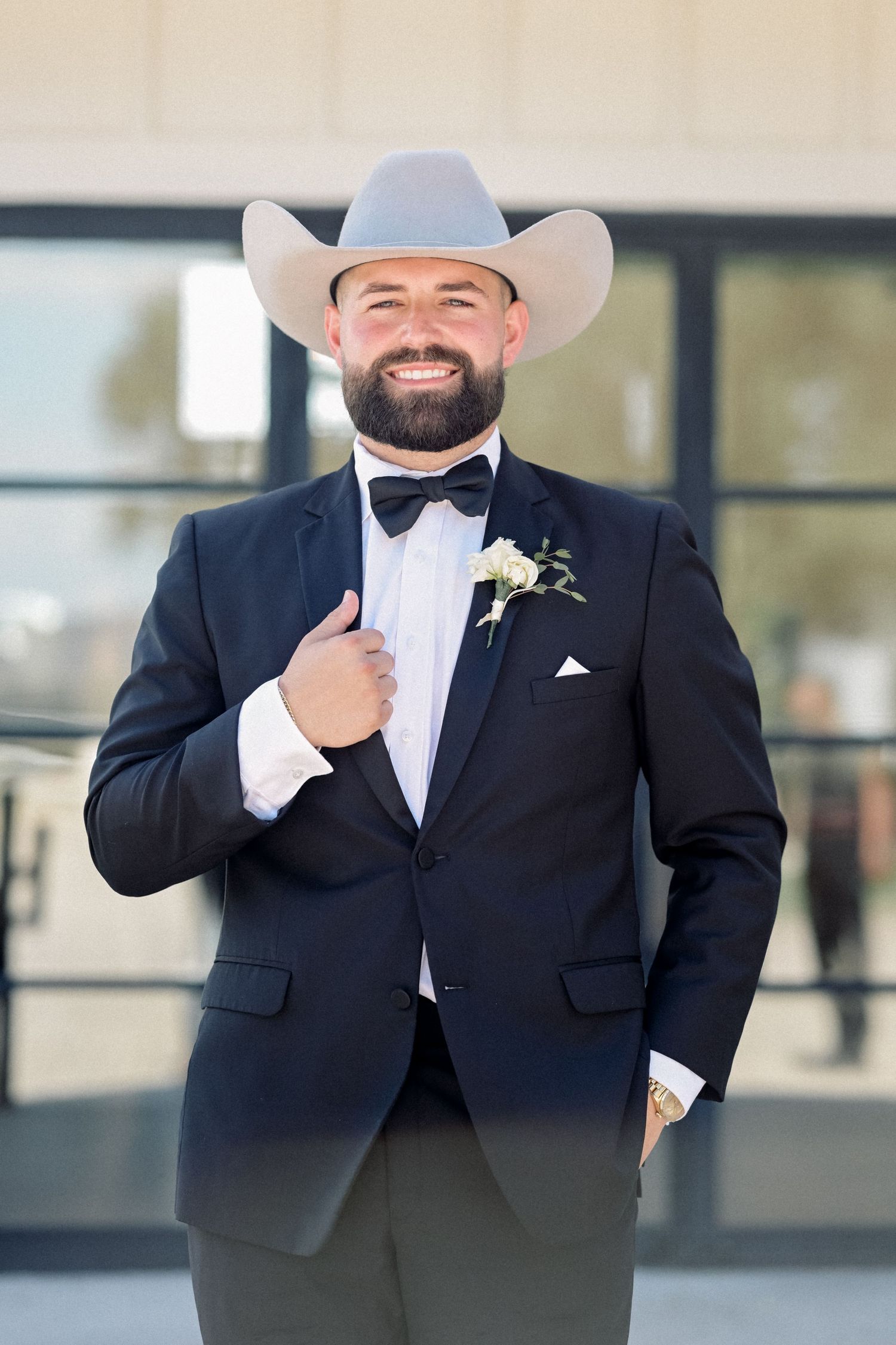 Smiling groom in black tuxedo and white cowboy hat with boutonniere poses outdoors on wedding day.