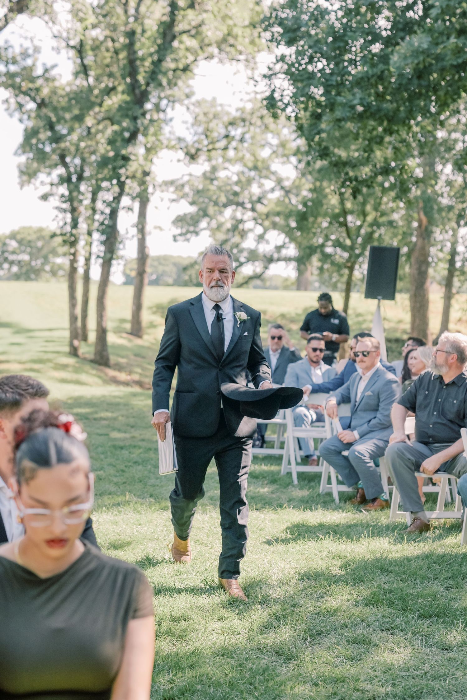 Older gentleman in dark suit walks down outdoor wedding aisle carrying a cowboy hat during ceremony.