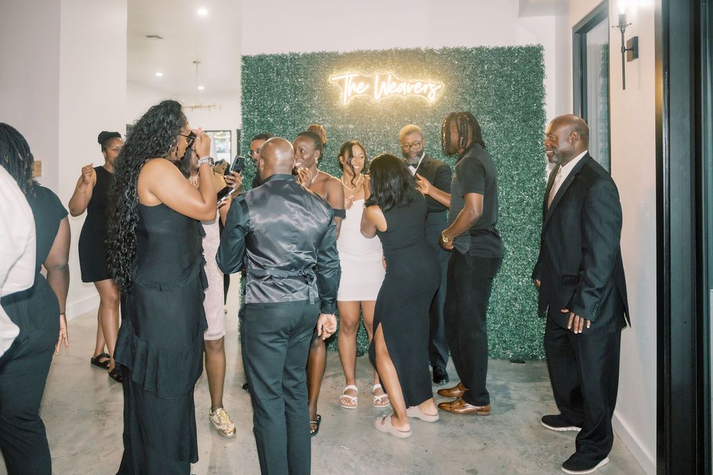 Guests in formal attire socializing at an elegant event near a green hedge wall with a neon sign.