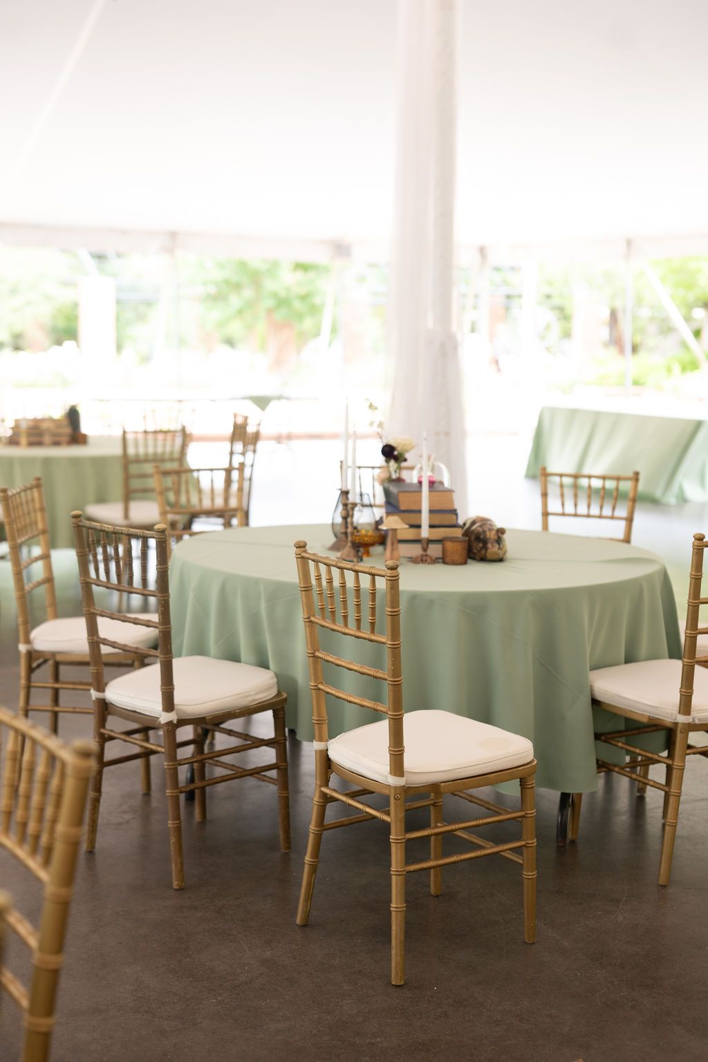 Elegant event tent with round tables draped in sage green linens and gold Chiavari chairs with white cushions.