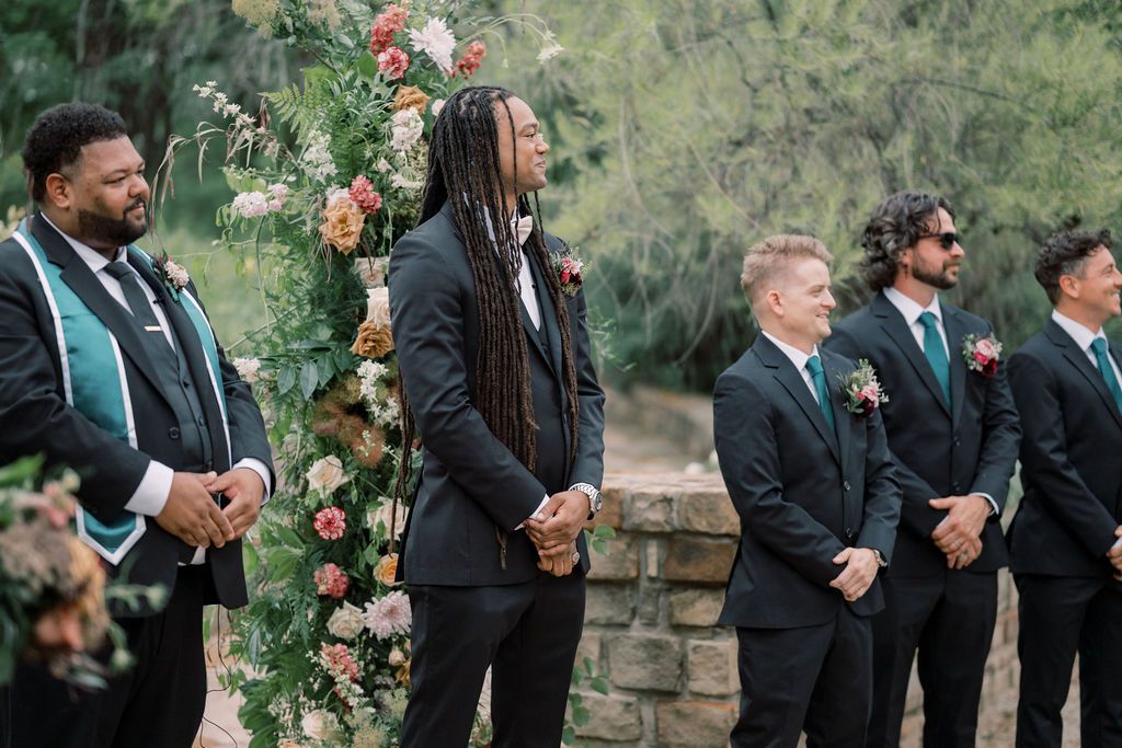 Groomsmen in black suits with green ties stand at outdoor wedding ceremony beside floral arch with pink and white blooms.