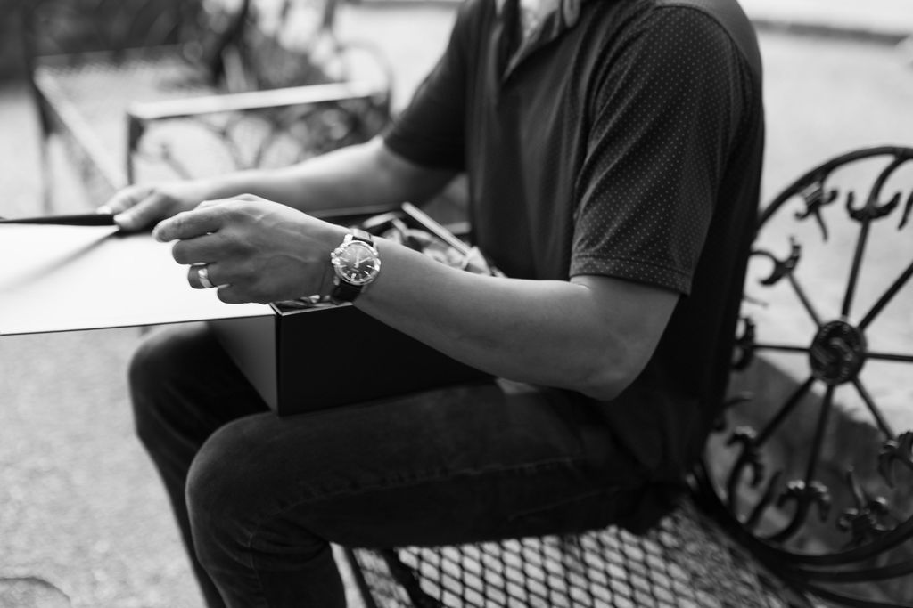 Man in black shirt wearing a watch sits on ornate bench, writing on paper in black and white photo.