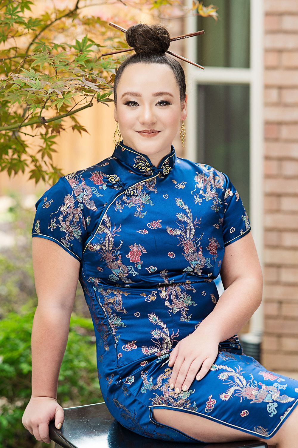 Woman in blue floral qipao dress with hair chopsticks poses outdoors near autumn foliage.