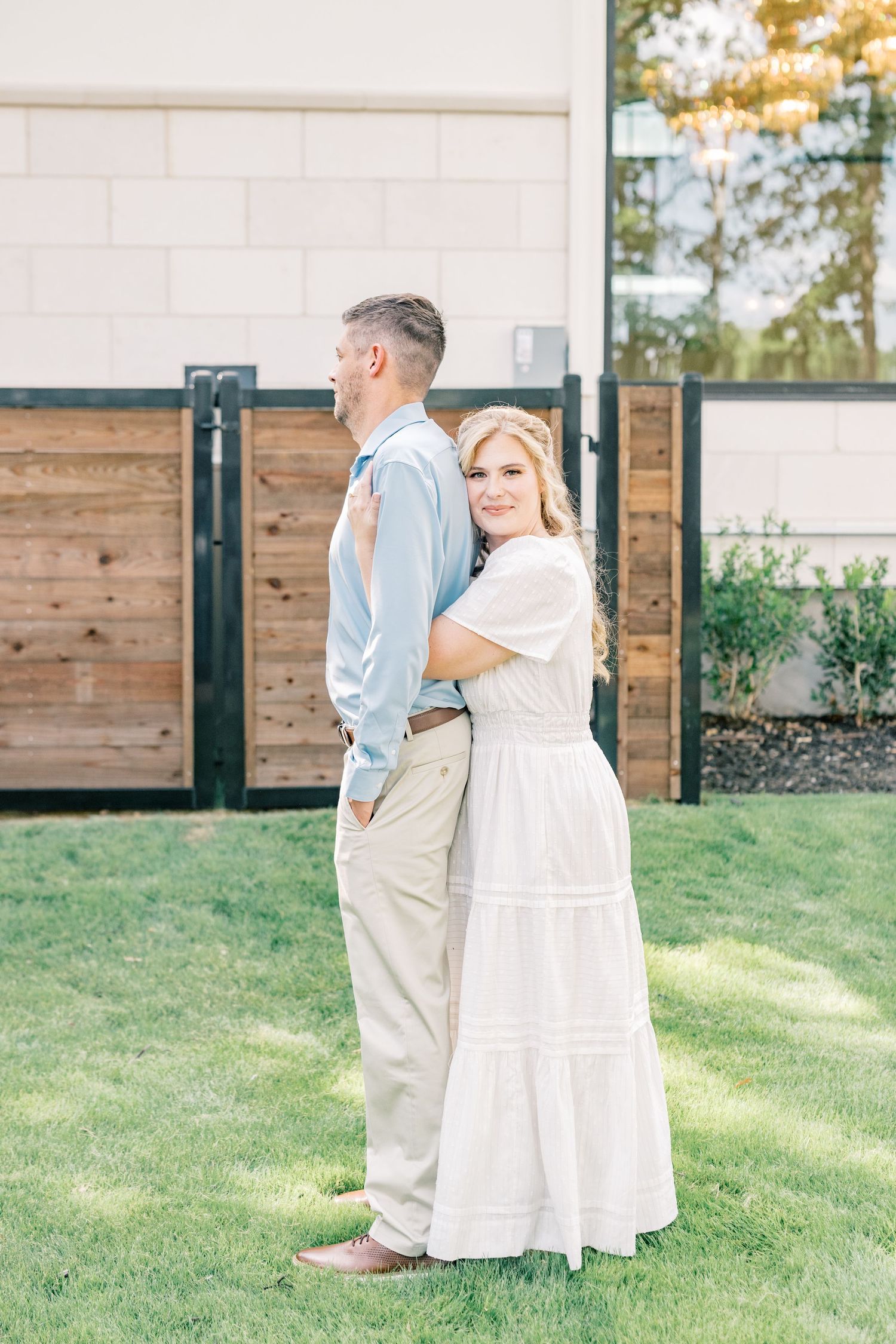 Couple posing back-to-back on green lawn, woman in white maxi dress hugging man in light blue shirt.