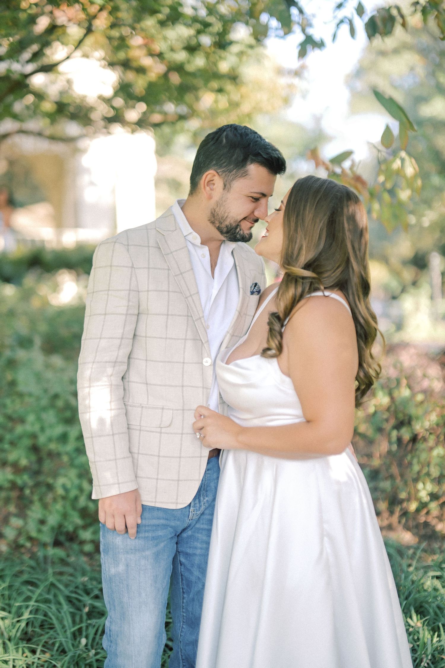 Engaged couple touch noses in a lush green park, she wearing a white dress and he a cream plaid blazer and jeans.