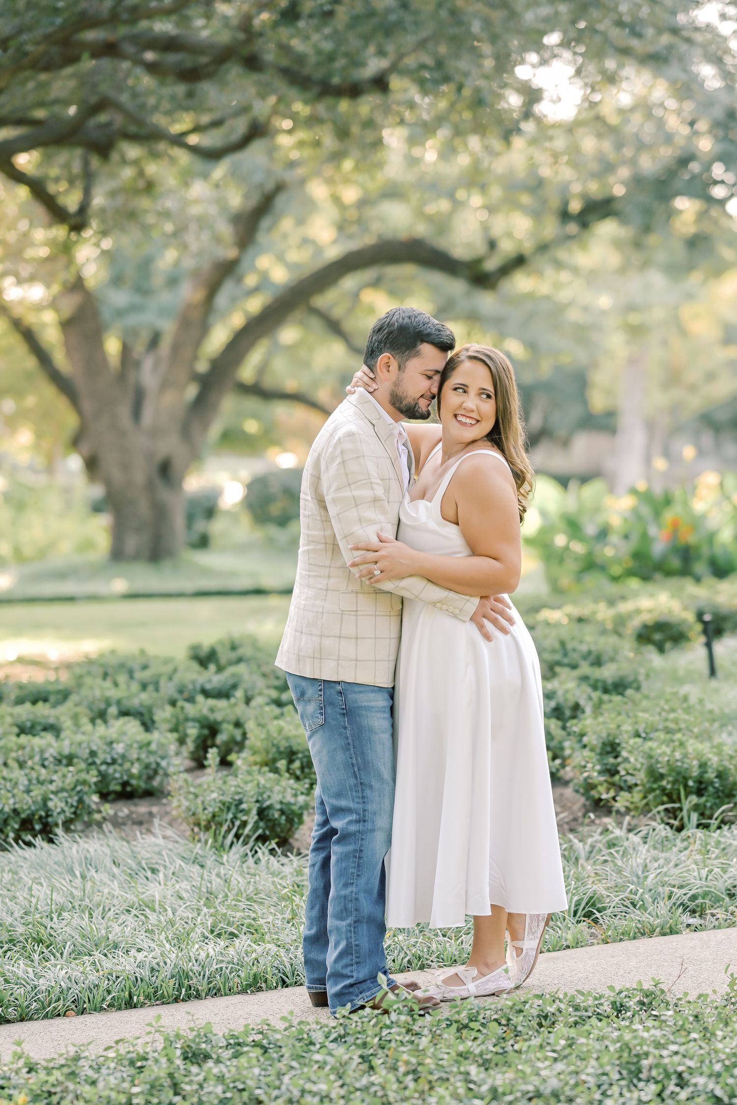 Smiling couple embracing on a garden path beneath large oak trees during an outdoor engagement photo session.