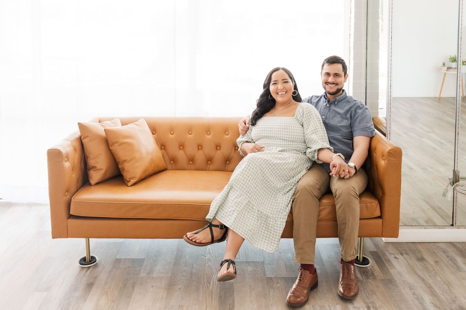 A couple sits casually together on a tan leather couch in a bright modern room with hardwood floors.