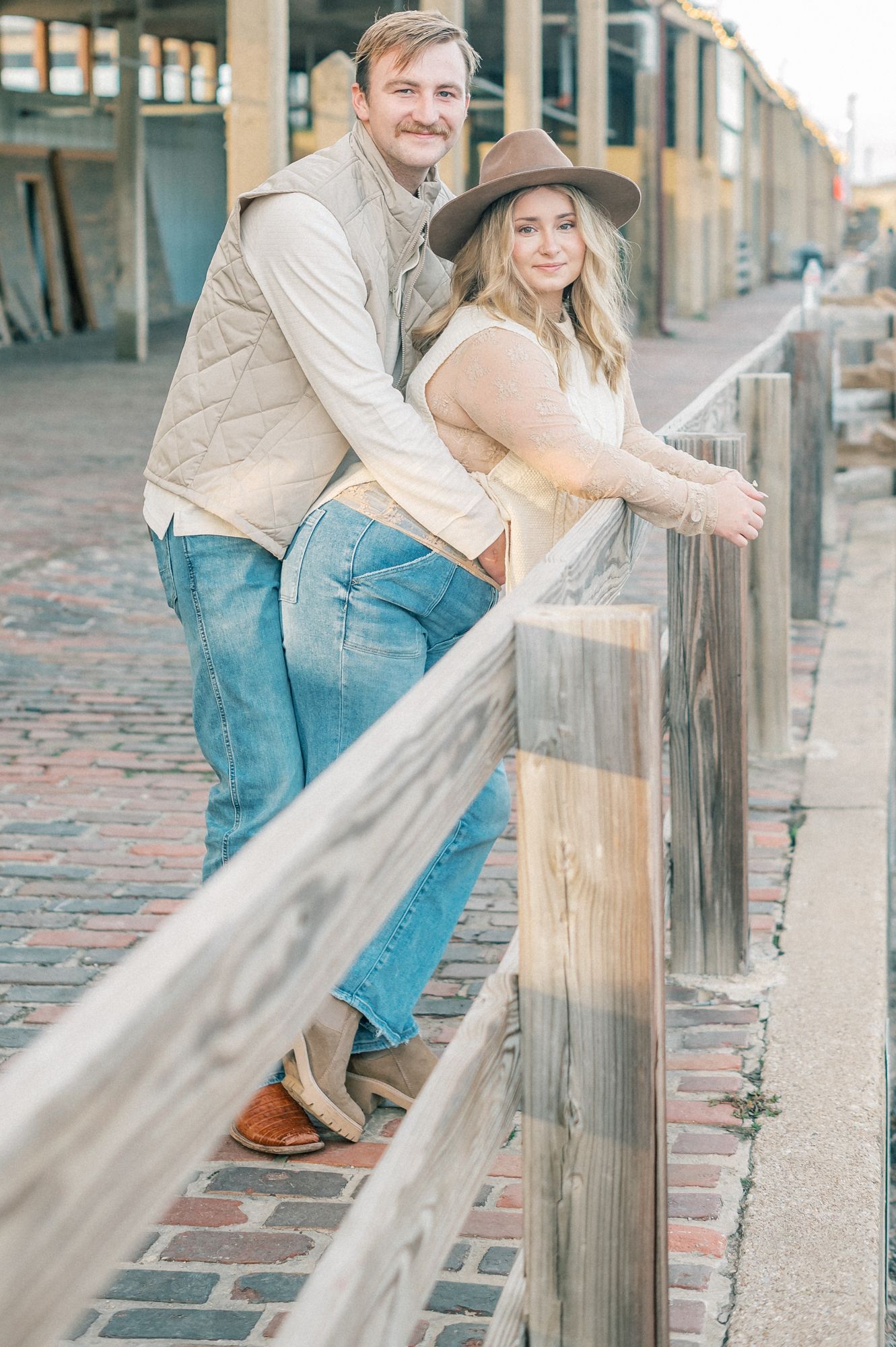A couple in western attire leans playfully against a wooden railing in an urban setting.