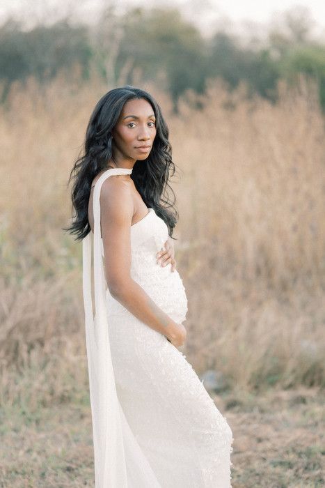 A bride in a flowing white dress stands in a sunlit field of tall grass during a romantic outdoor photoshoot.